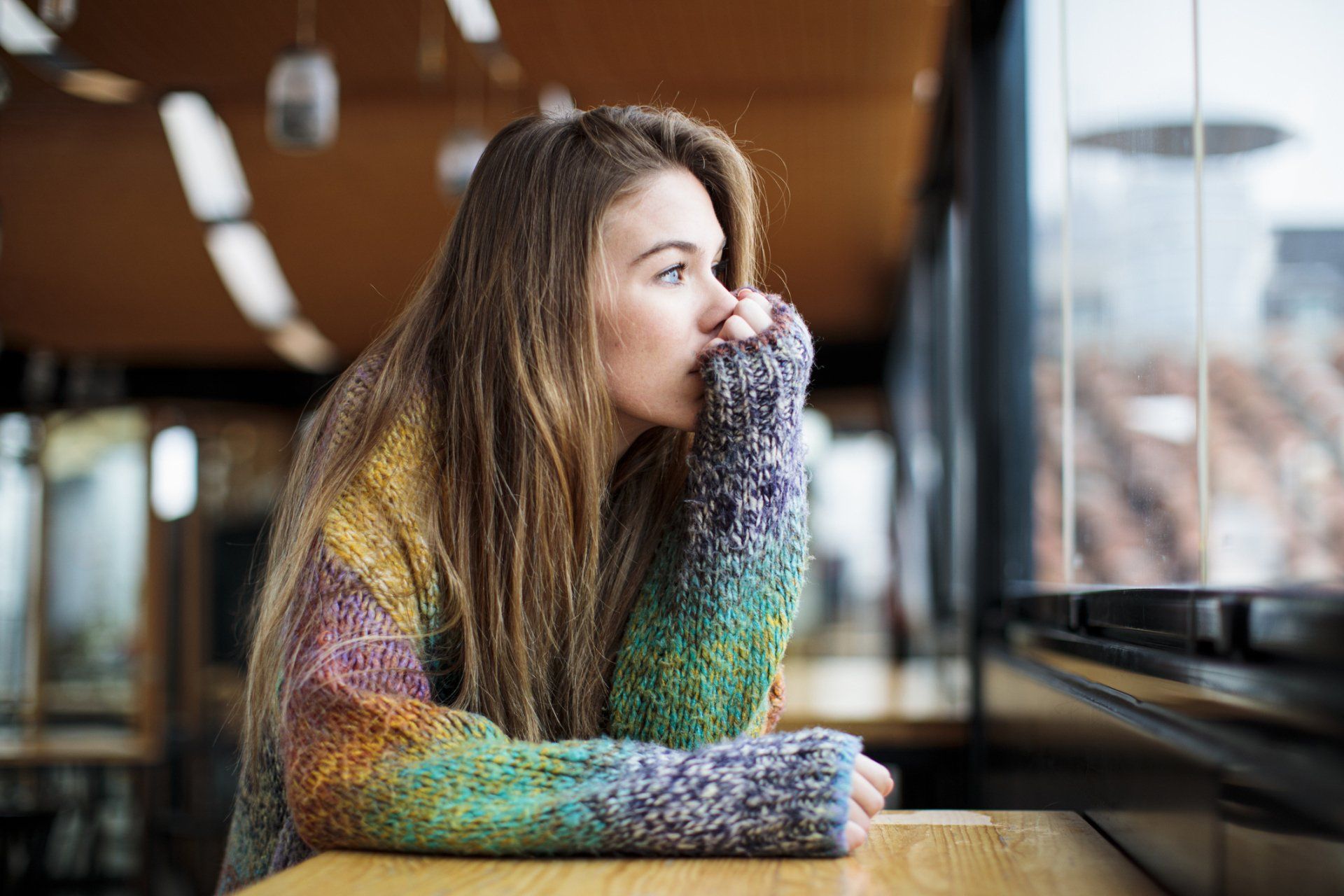 Woman looking out window, resting chin on hand, wearing colorful sweater in a cafe setting.