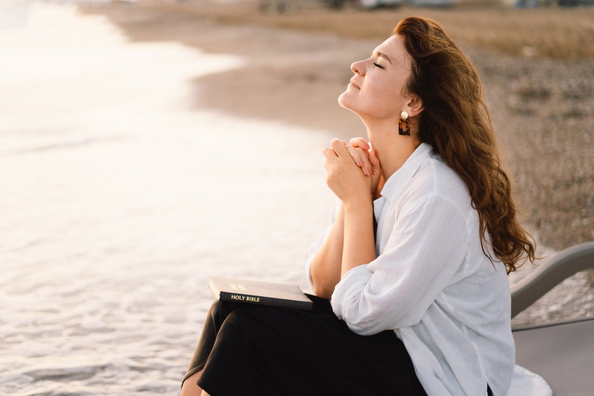 Woman praying with hands clasped, eyes closed, near the ocean.