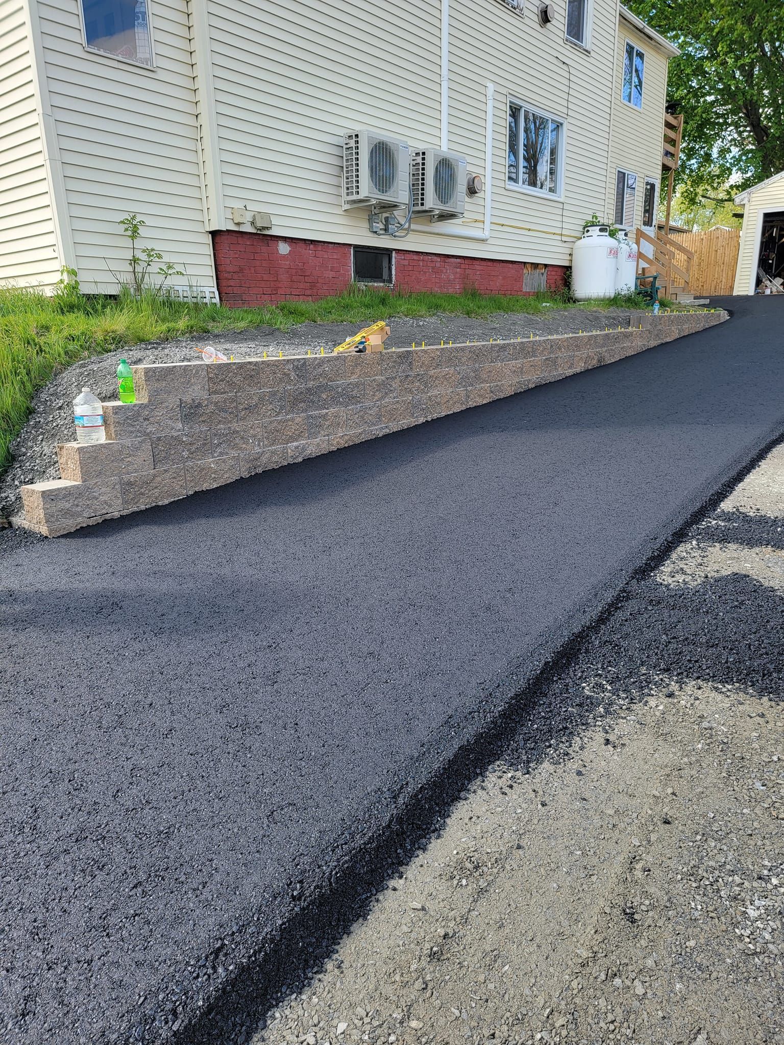 A newly paved asphalt driveway borders a stepped stone retaining wall next to a light-colored house with AC units.