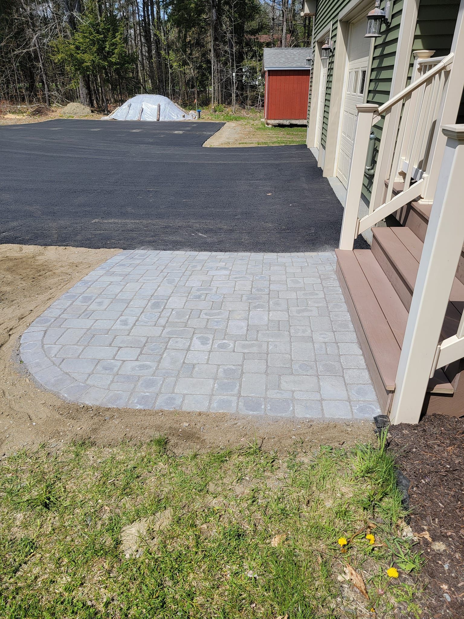 A gray stone paver landing at the base of wooden stairs, adjacent to a black paved driveway and a green building.