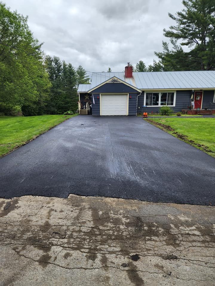 Asphalt driveway between brick building and gray-sided house. Wet surface, sidewalk in foreground.