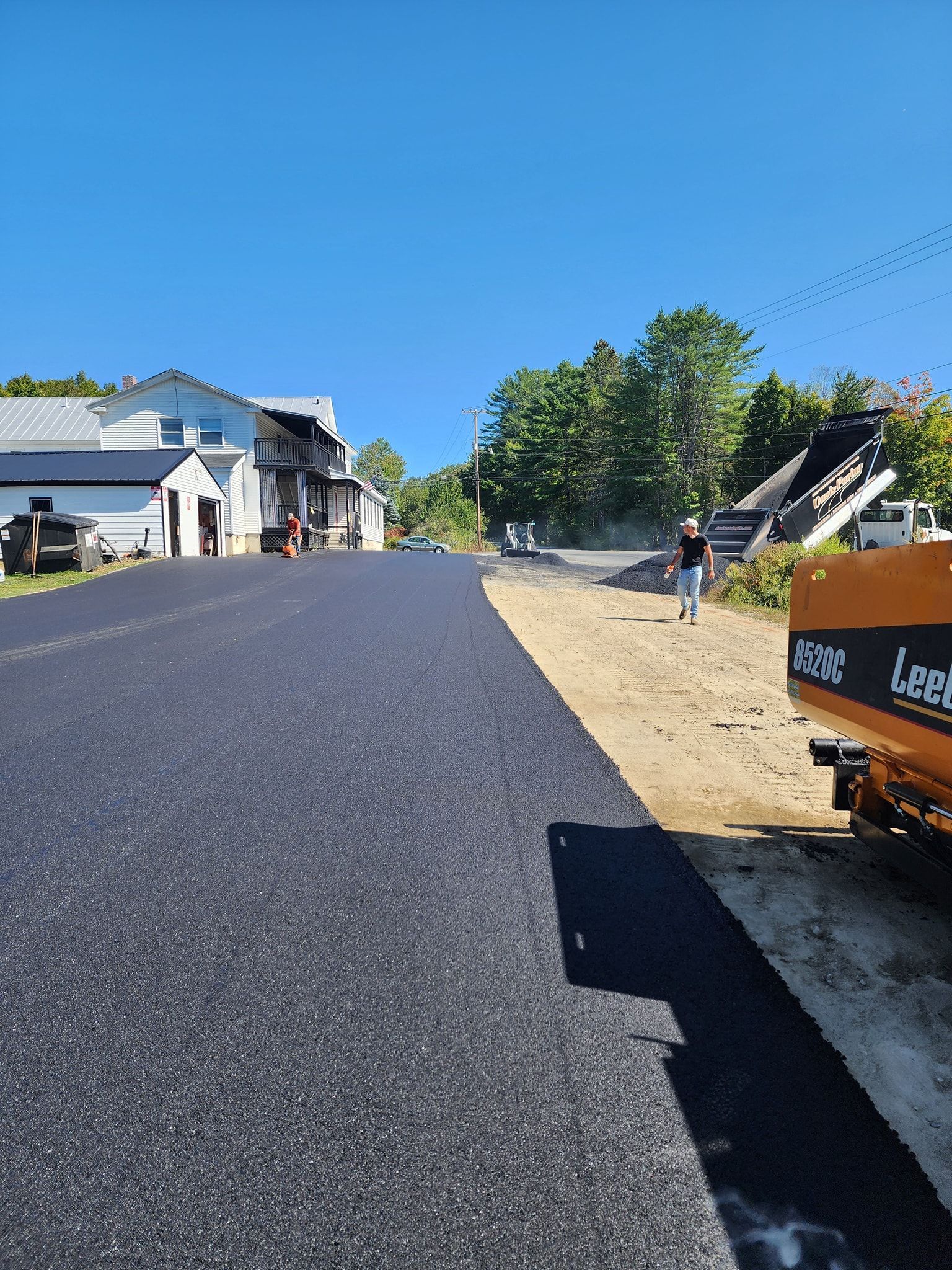 Fresh asphalt driveway at a construction site with a yellow paving machine and people walking nearby on a sunny day.