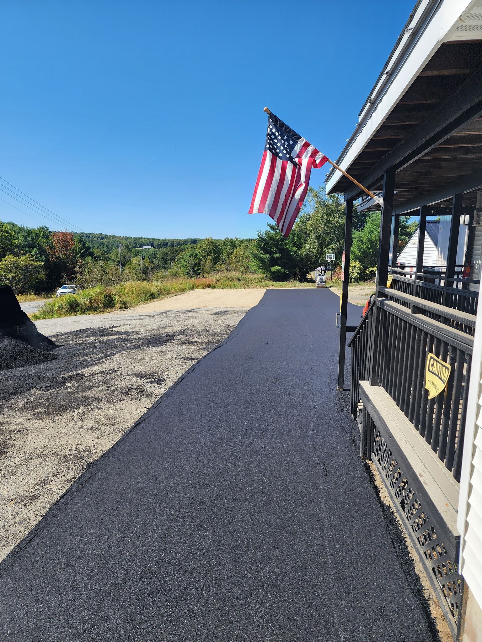 A freshly paved black asphalt walkway runs alongside a house porch with an American flag flying on a sunny day.