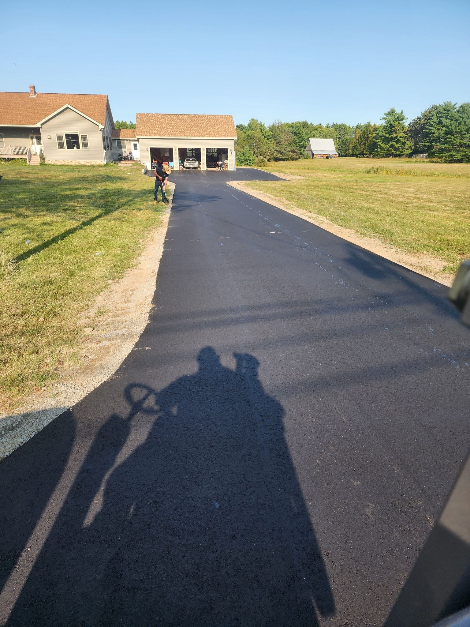 A newly paved black asphalt driveway leads toward a house with an attached garage under a clear blue sky.