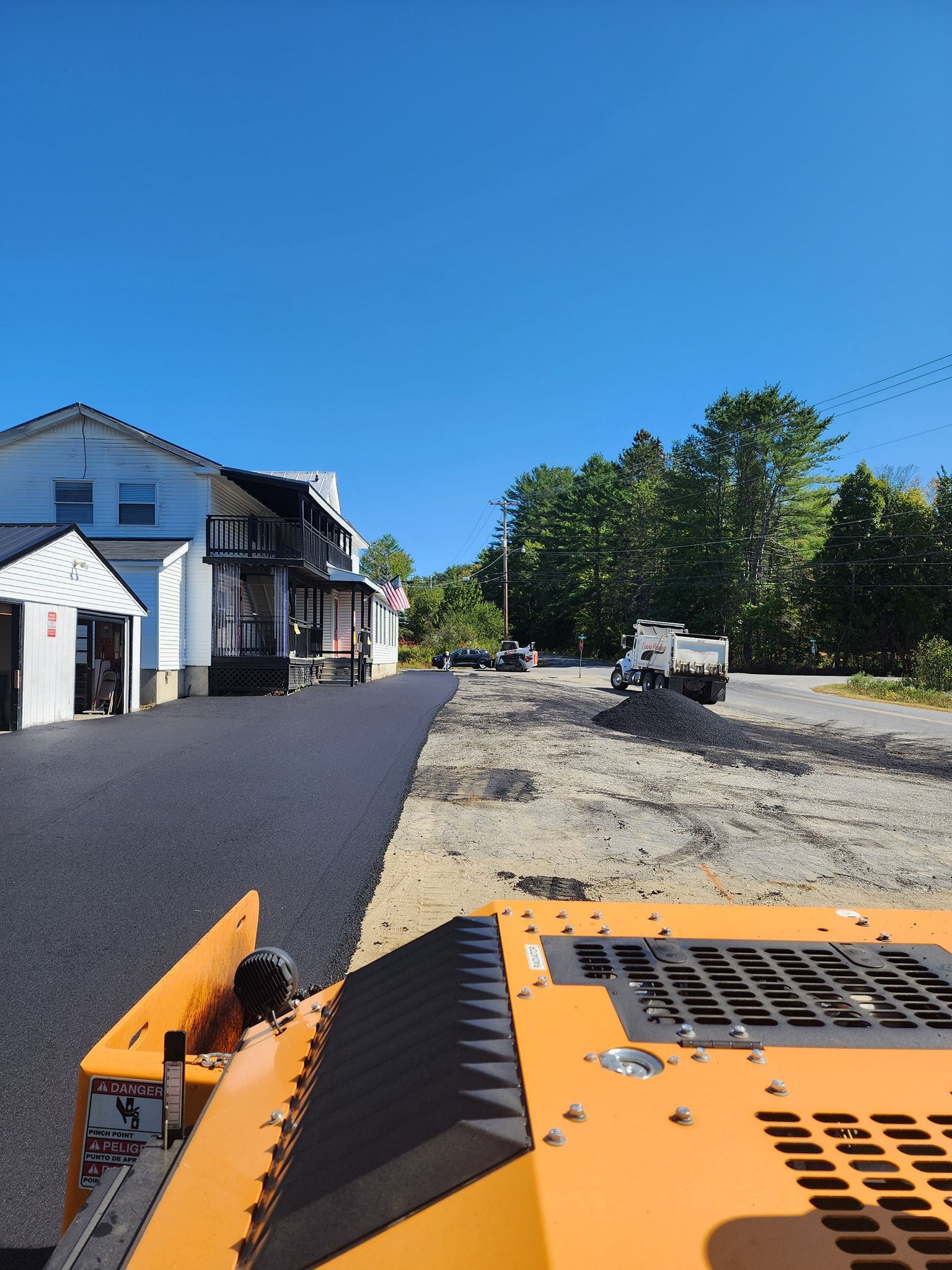 View from a paving machine showing a newly laid asphalt driveway beside a white building and a gravel construction area.