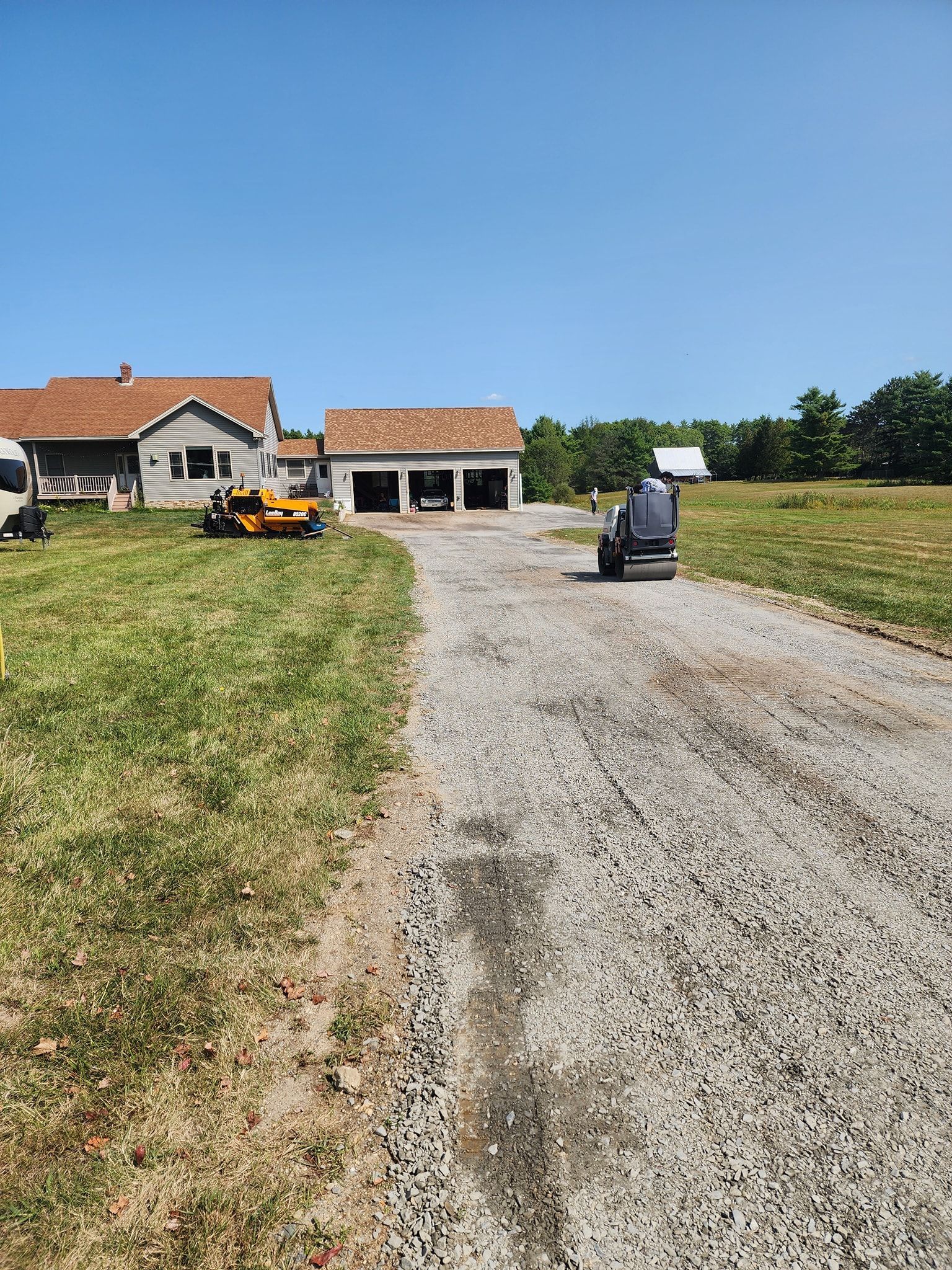 A gravel driveway leads toward a house and a multi-car garage on a sunny day with mowers parked on the lawn.