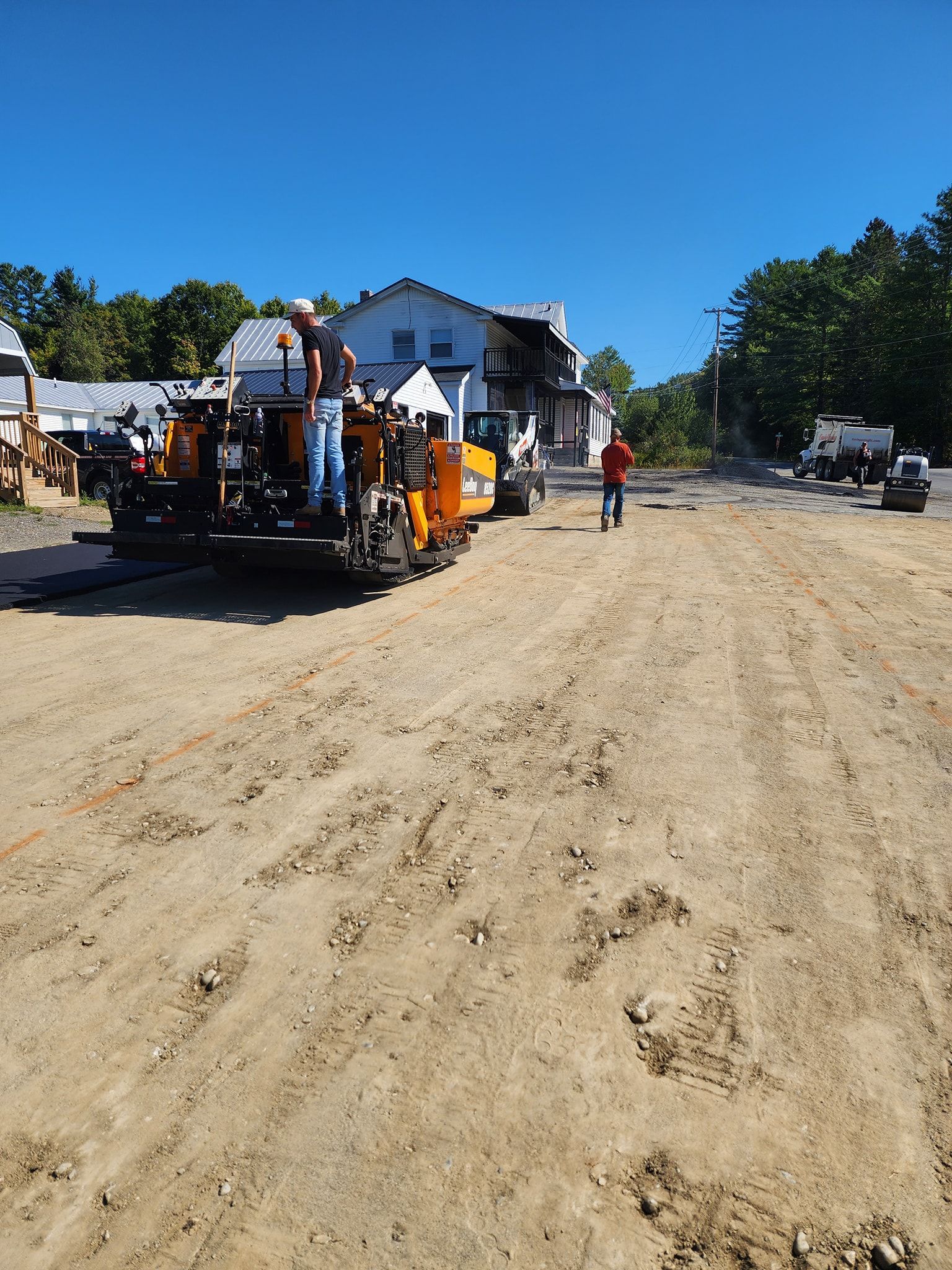Construction workers operate a yellow paving machine on a gravel lot near a white building under a clear blue sky.