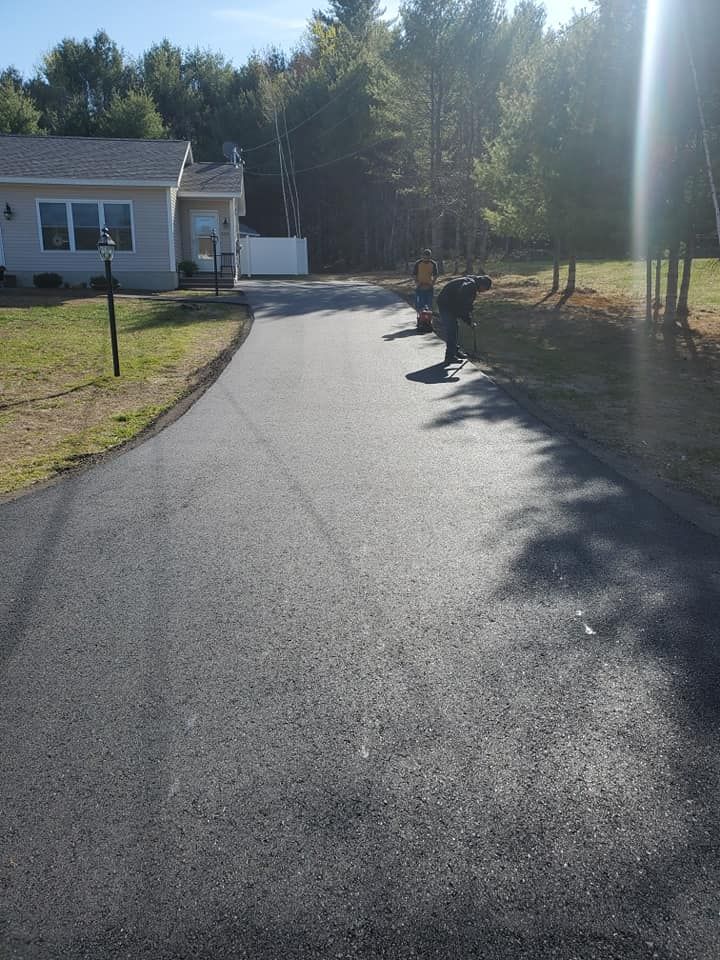 Workers use tools to finish the edges of a newly paved asphalt driveway next to a house on a sunny day.