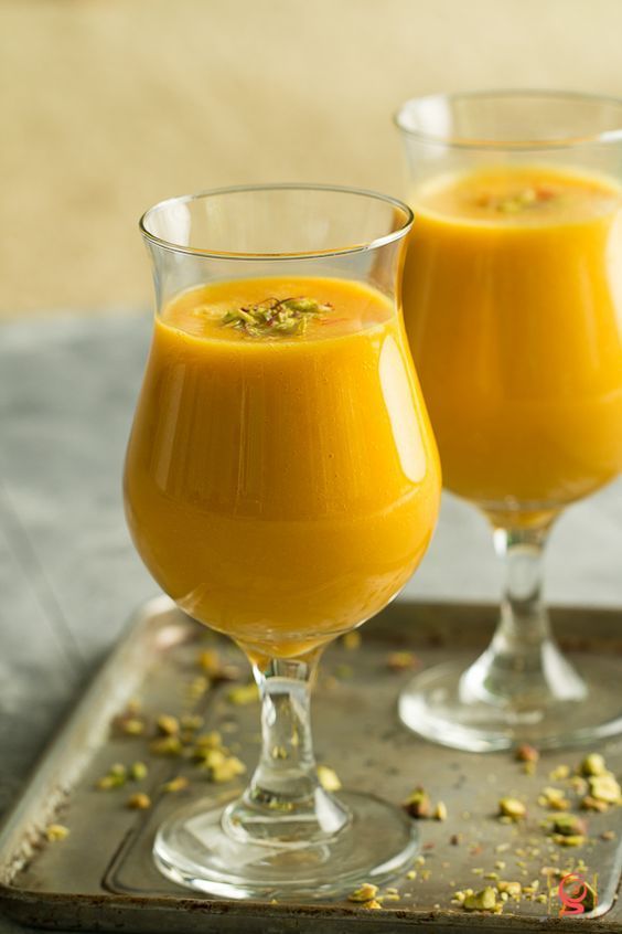 Two mango lassi drinks in stemmed glasses, on a silver tray, sprinkled with pistachios.