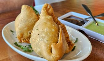 Two golden samosas on a white plate, served with dark and green chutneys.
