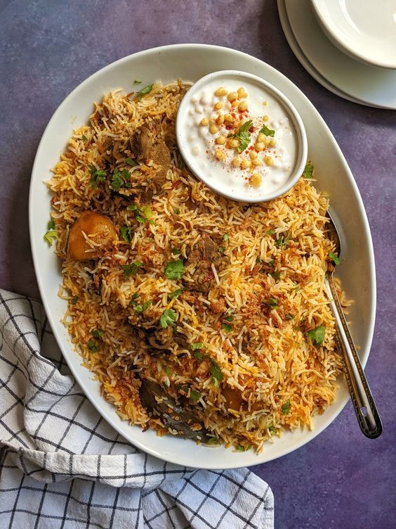 Oval platter of biryani with raita, garnished with cilantro, on a checkered napkin.