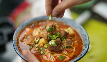 Person sprinkles chopped green onions onto a bowl of beef noodle soup.