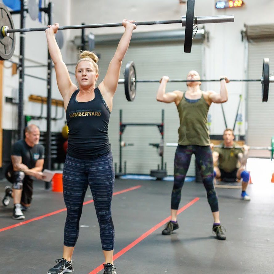 A woman is lifting a barbell in a gym while a man sits in the background