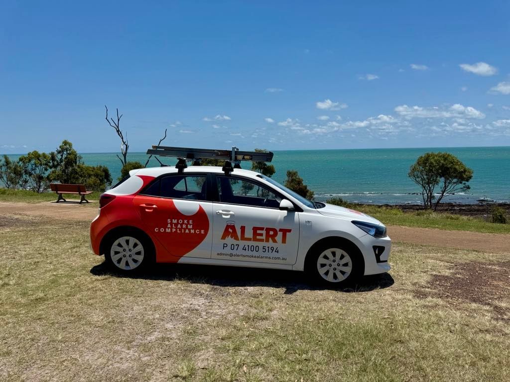 A White and Red Alert Car is Parked in Front of the Ocean — Alert Smoke Alarm Compliance in Maryborough, QLD