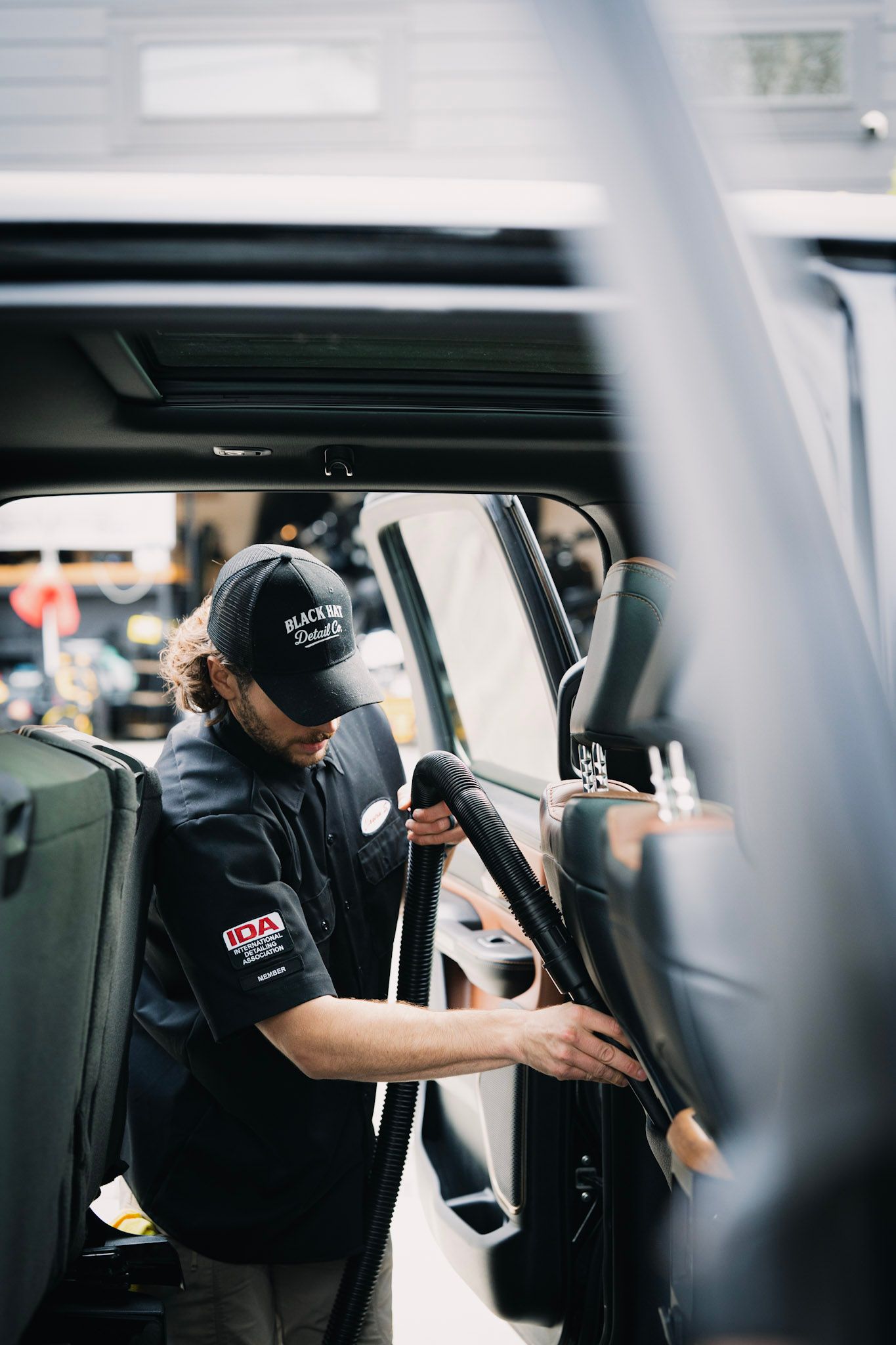 Person adjusts the seat inside a vehicle. Black shirt, cap, focused expression. Bright setting.