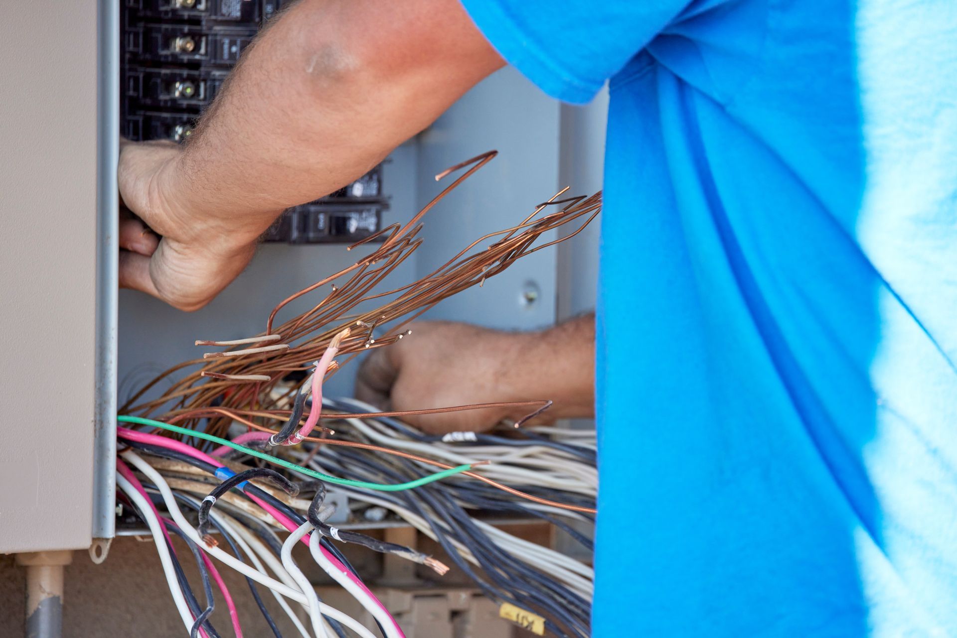 Person in a blue shirt working on electrical wiring inside a gray electrical panel box outdoors.