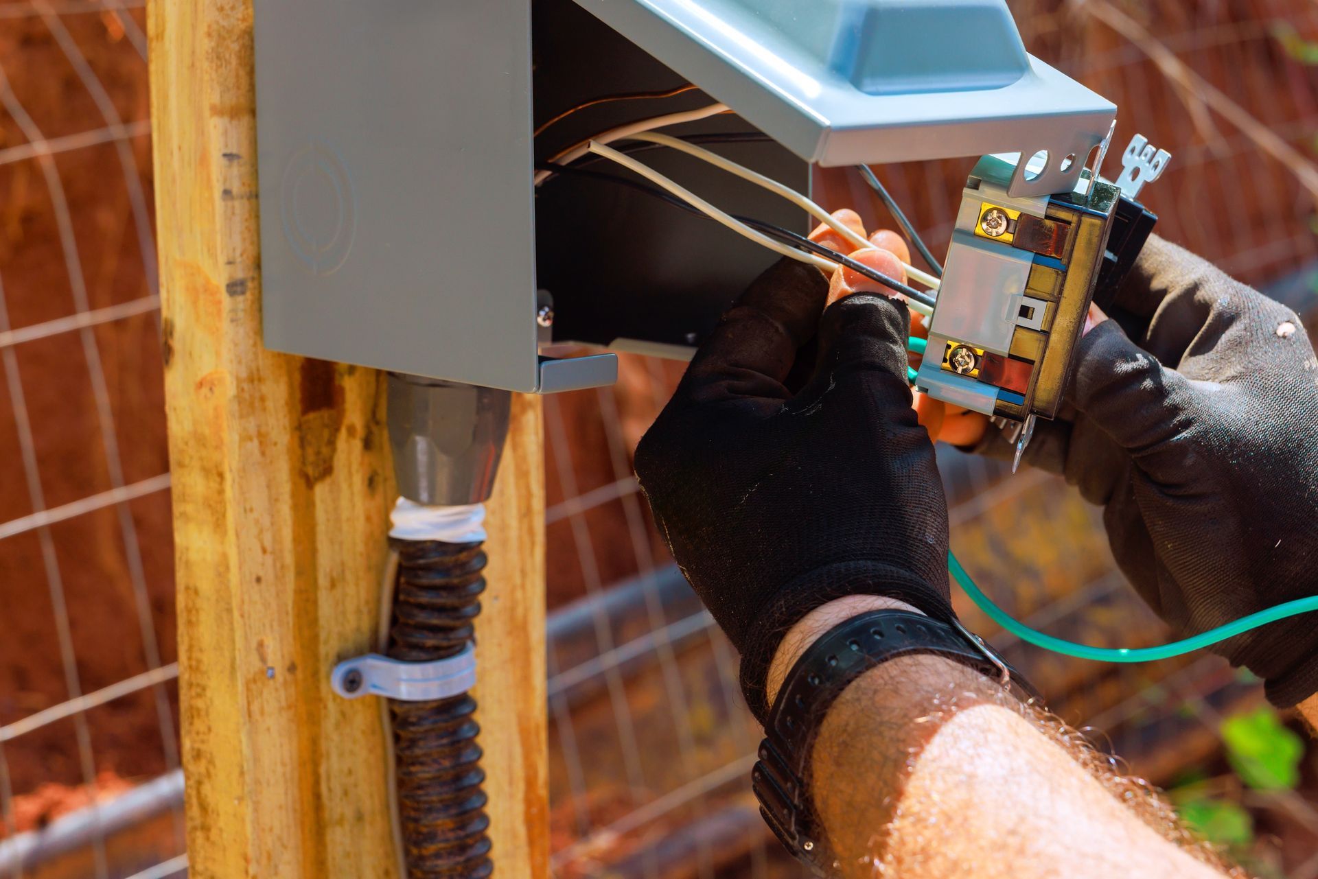 Person in black gloves wiring an electrical outlet in a gray box, attached to a wooden post outside.