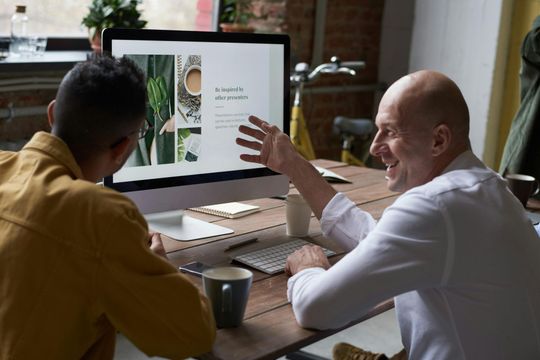 Two men are sitting at a table looking at a computer screen.