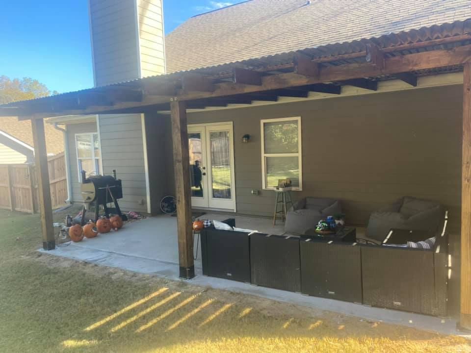 A covered patio with outdoor furniture and a grill sits next to a house with a clear blue sky above.