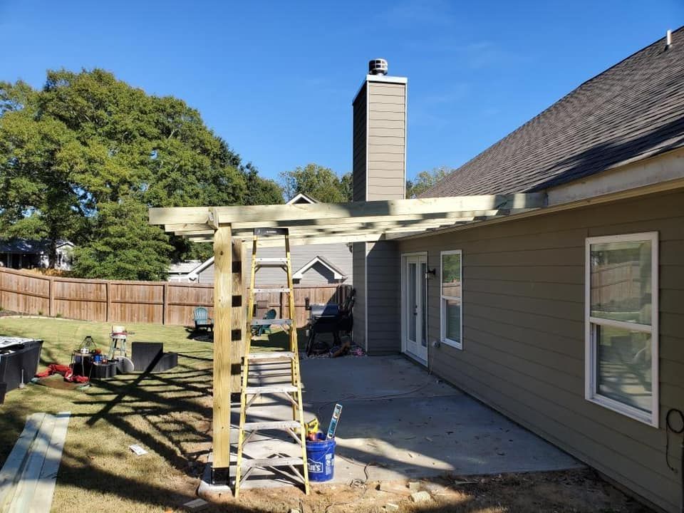 Wooden pergola construction in a backyard, beside a house with a chimney and patio.
