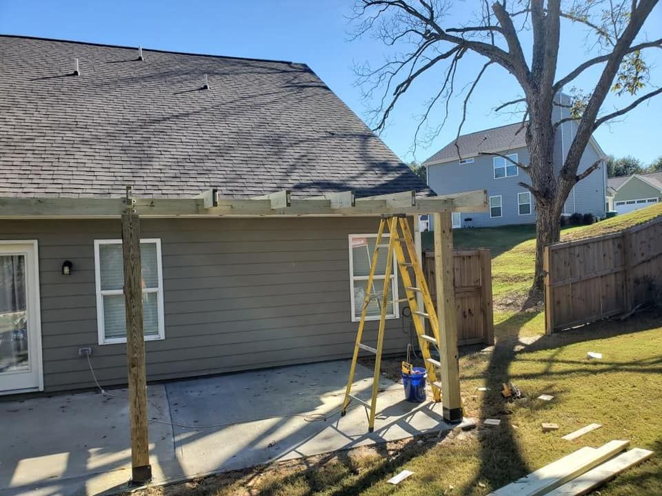 Construction of a pergola attached to a house. Yellow ladder, wooden beams, green siding, and blue sky.