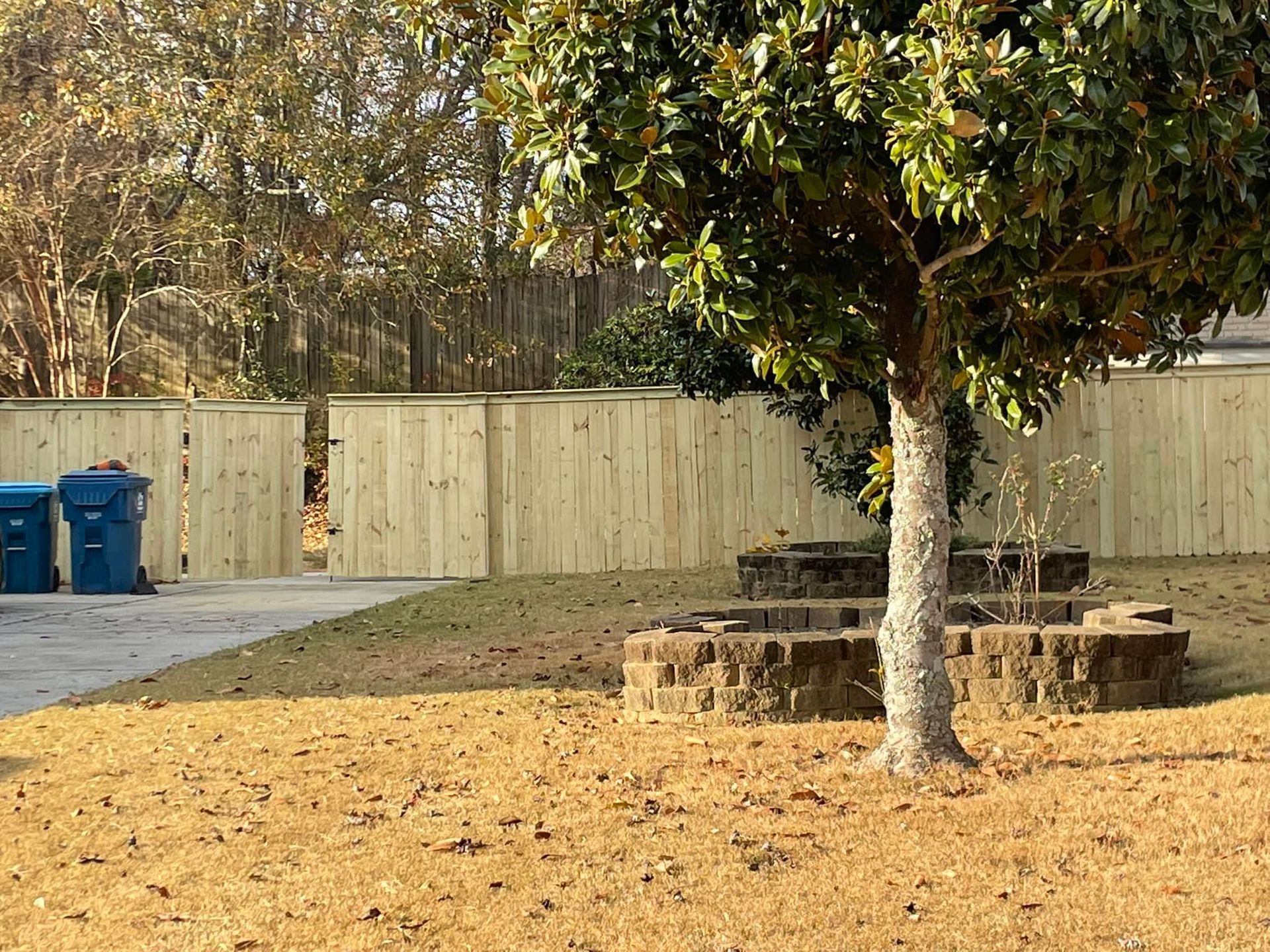 Backyard with a light-colored wooden fence, brown grass, and a tree with a brick-lined bed. Blue trash cans visible.