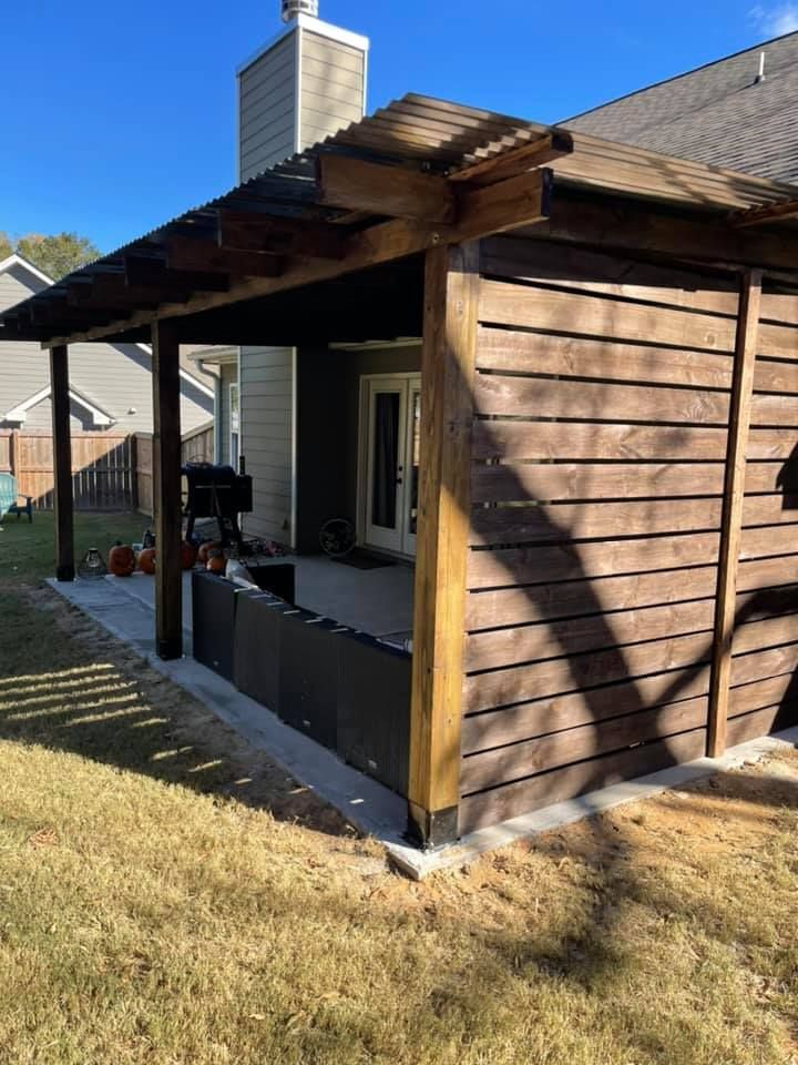 Outdoor wooden patio with a pergola and slatted wall, built next to a house with a green lawn.