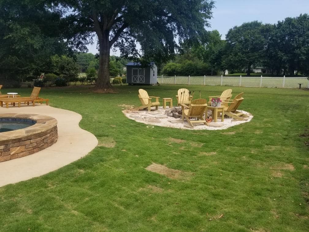 Lawn with fire pit and chairs. Stone pool surround on the left. Trees and small shed in the background.