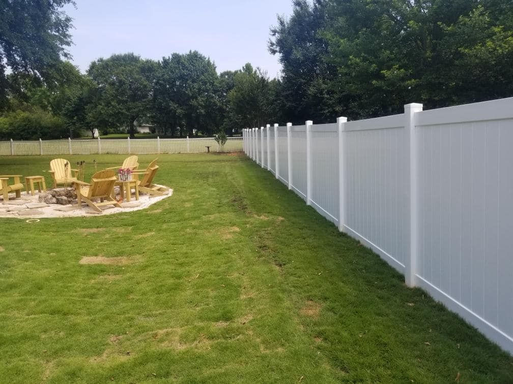 White vinyl fence in a grassy yard, fire pit and chairs visible in the background, trees under a blue sky.