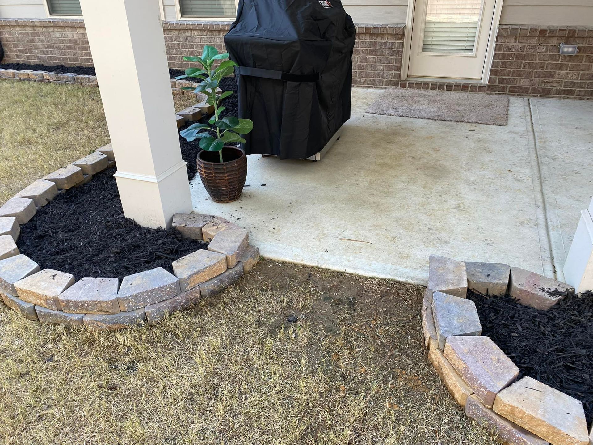 Patio with decorative brick borders, mulch, a grill, a potted plant, and a pillar.
