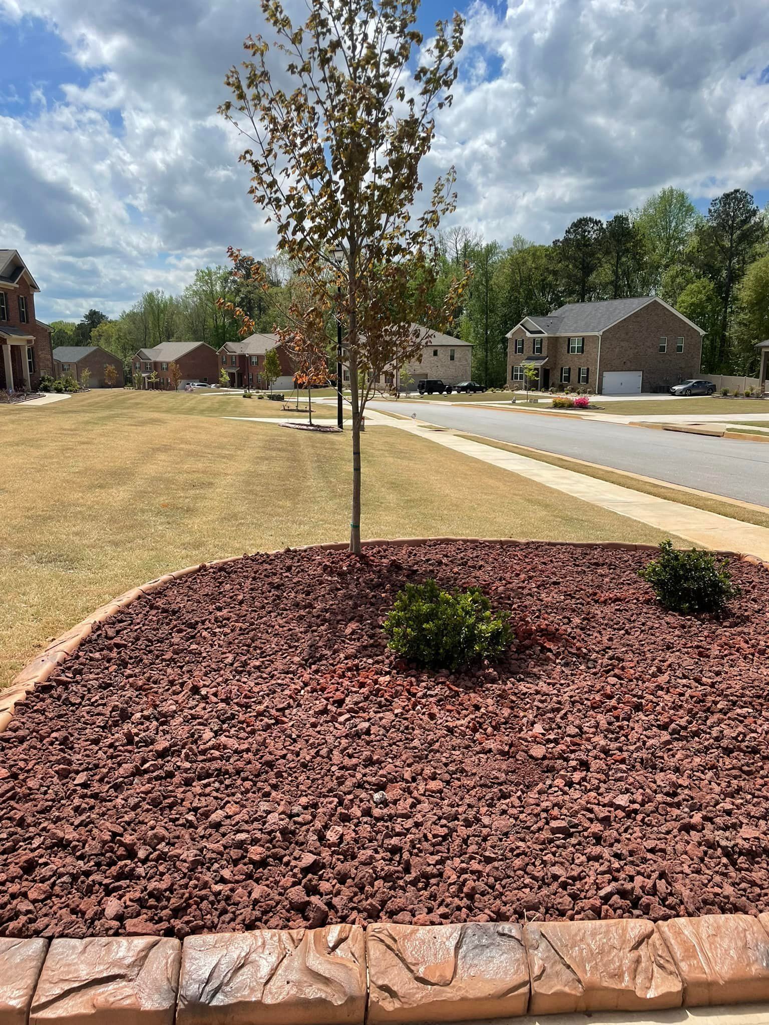 Red mulch bed with a tree and bushes, bordered by stone. Houses and street in the background.