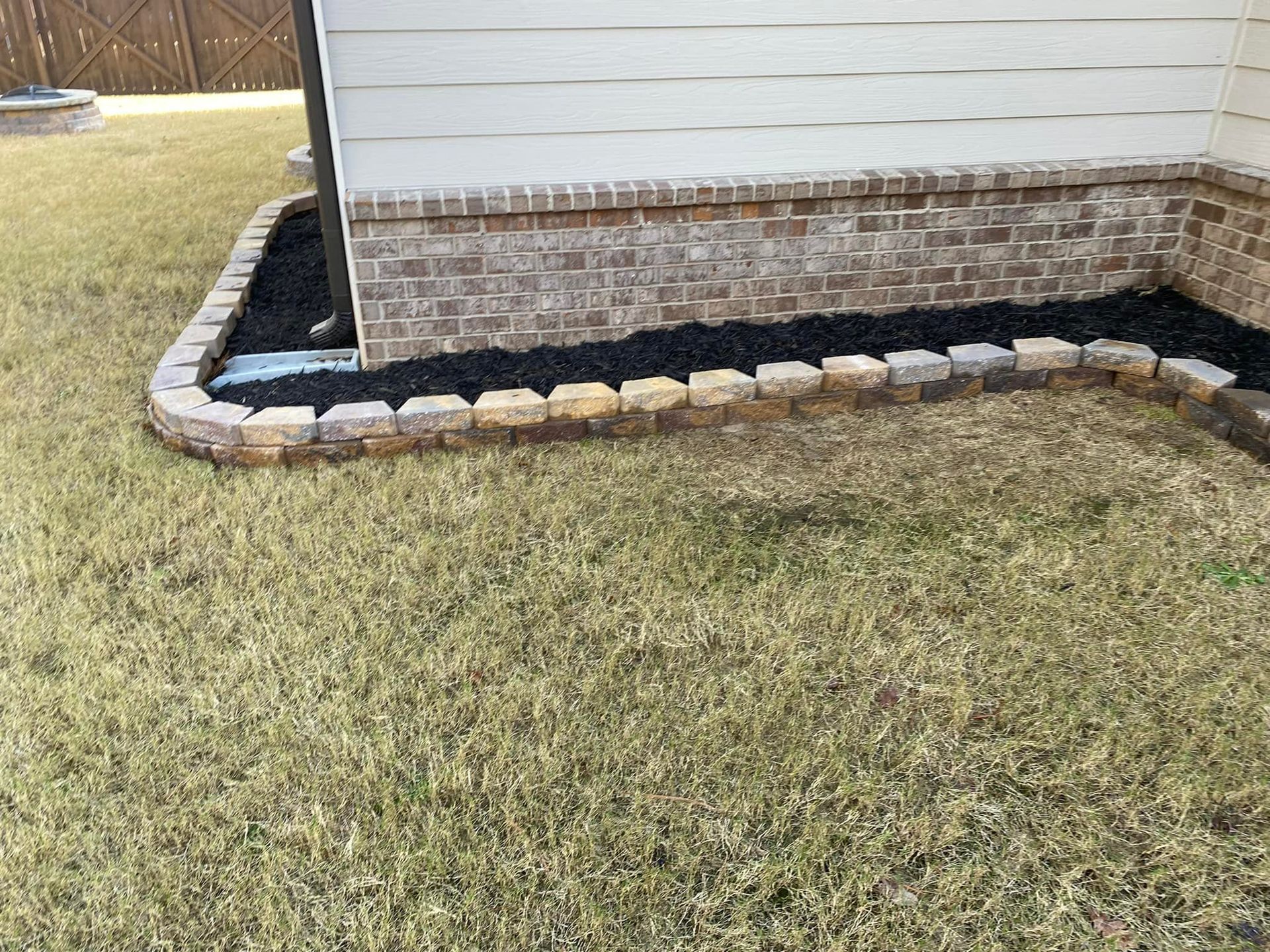 A low brick border encloses a bed of black mulch next to a building with brick foundation.