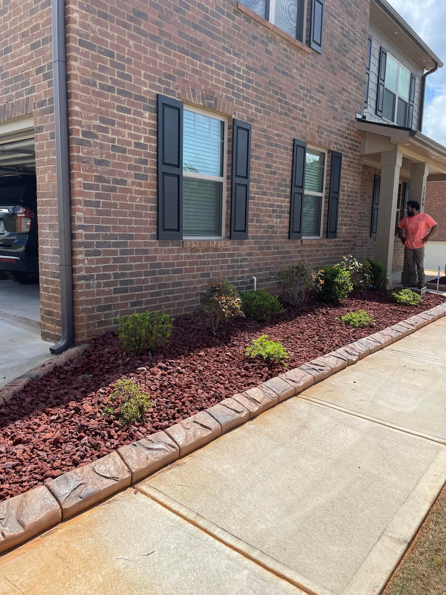 Brick house with dark shutters, red mulch landscaping, and a concrete walkway. Man stands on porch.