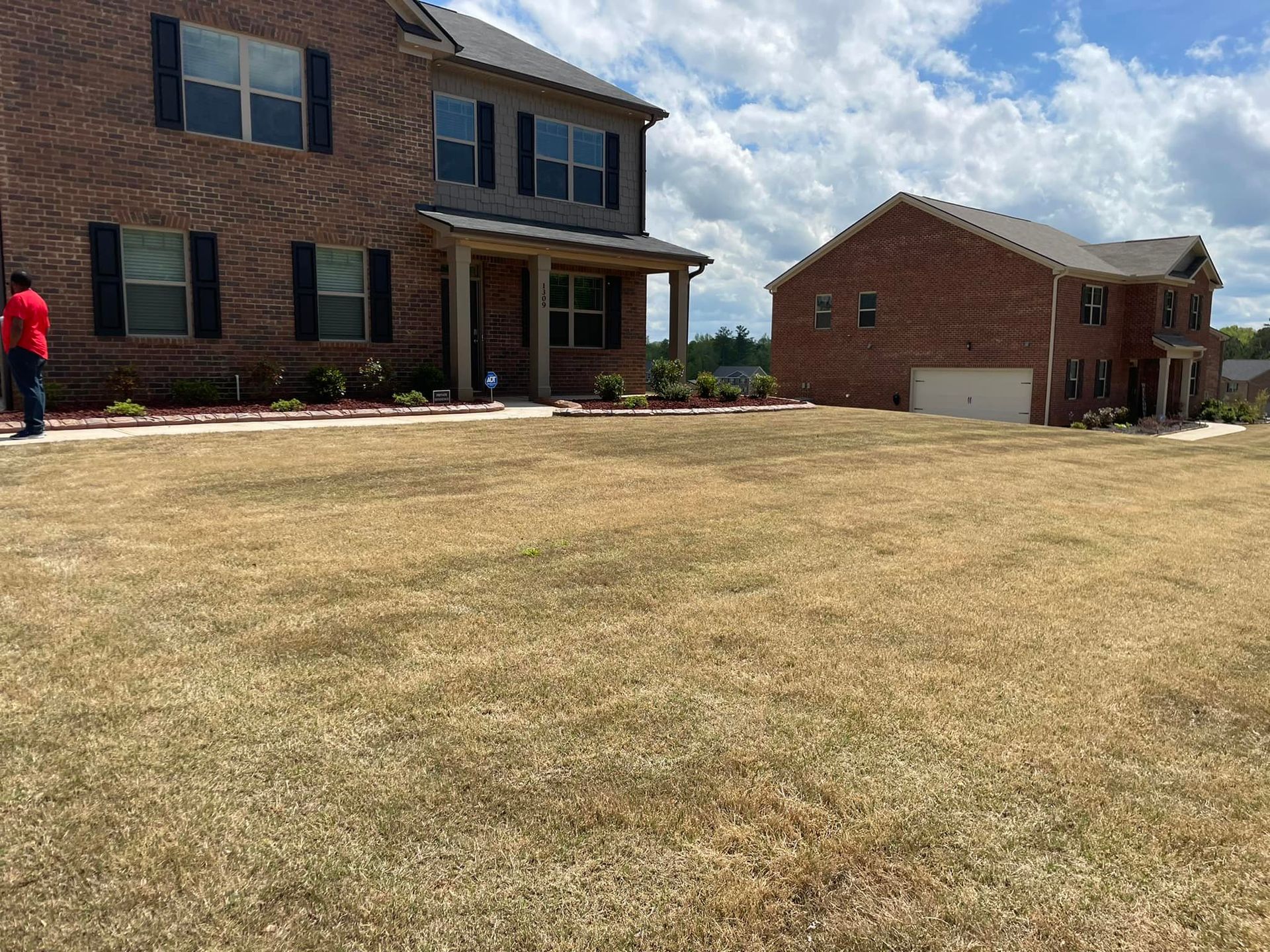 Brown, dead grass in front of a two-story brick house with dark shutters and a smaller brick building.