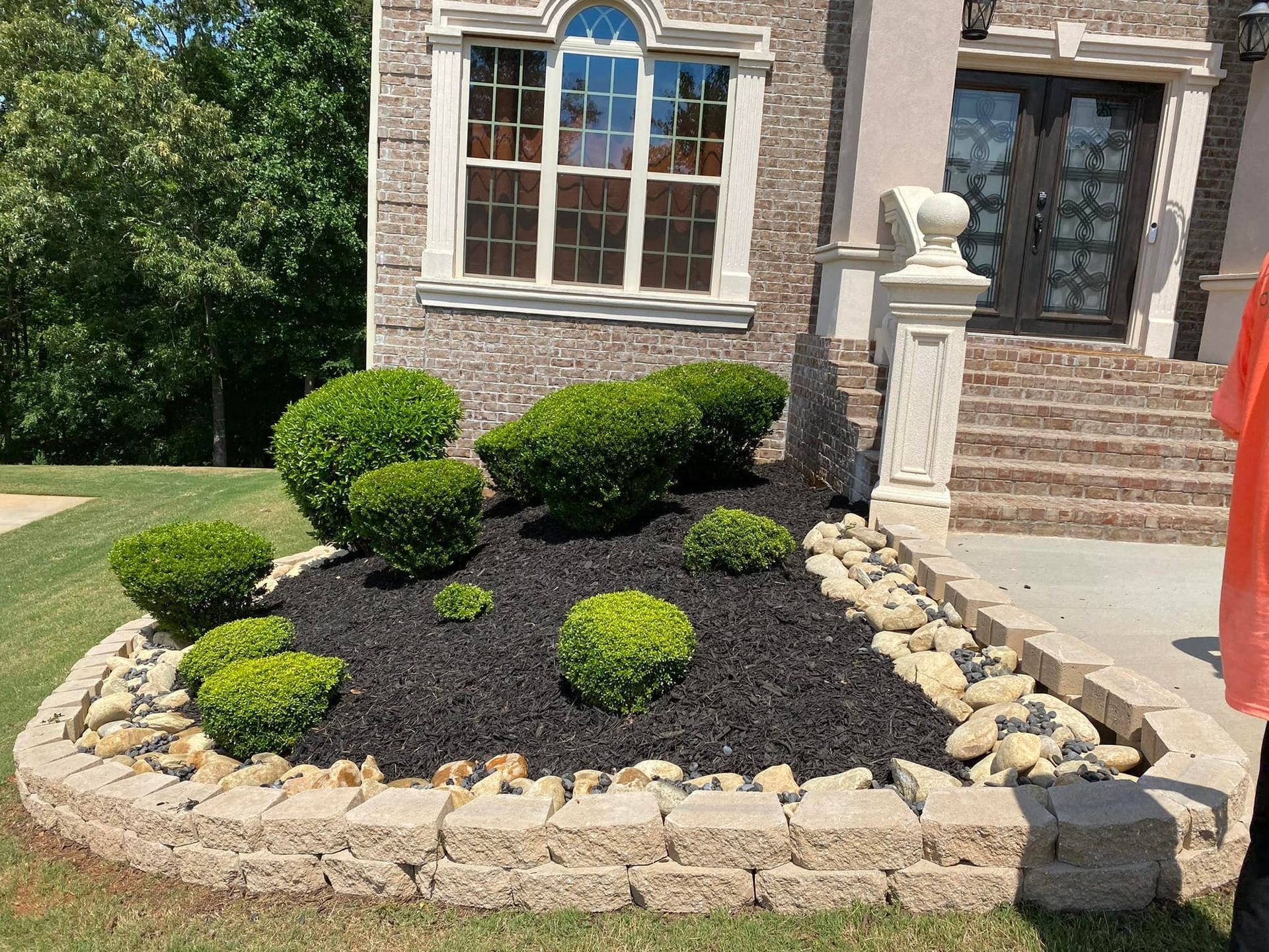 Landscaped front yard with a raised bed of green shrubs and black mulch, bordered by light-colored stones.