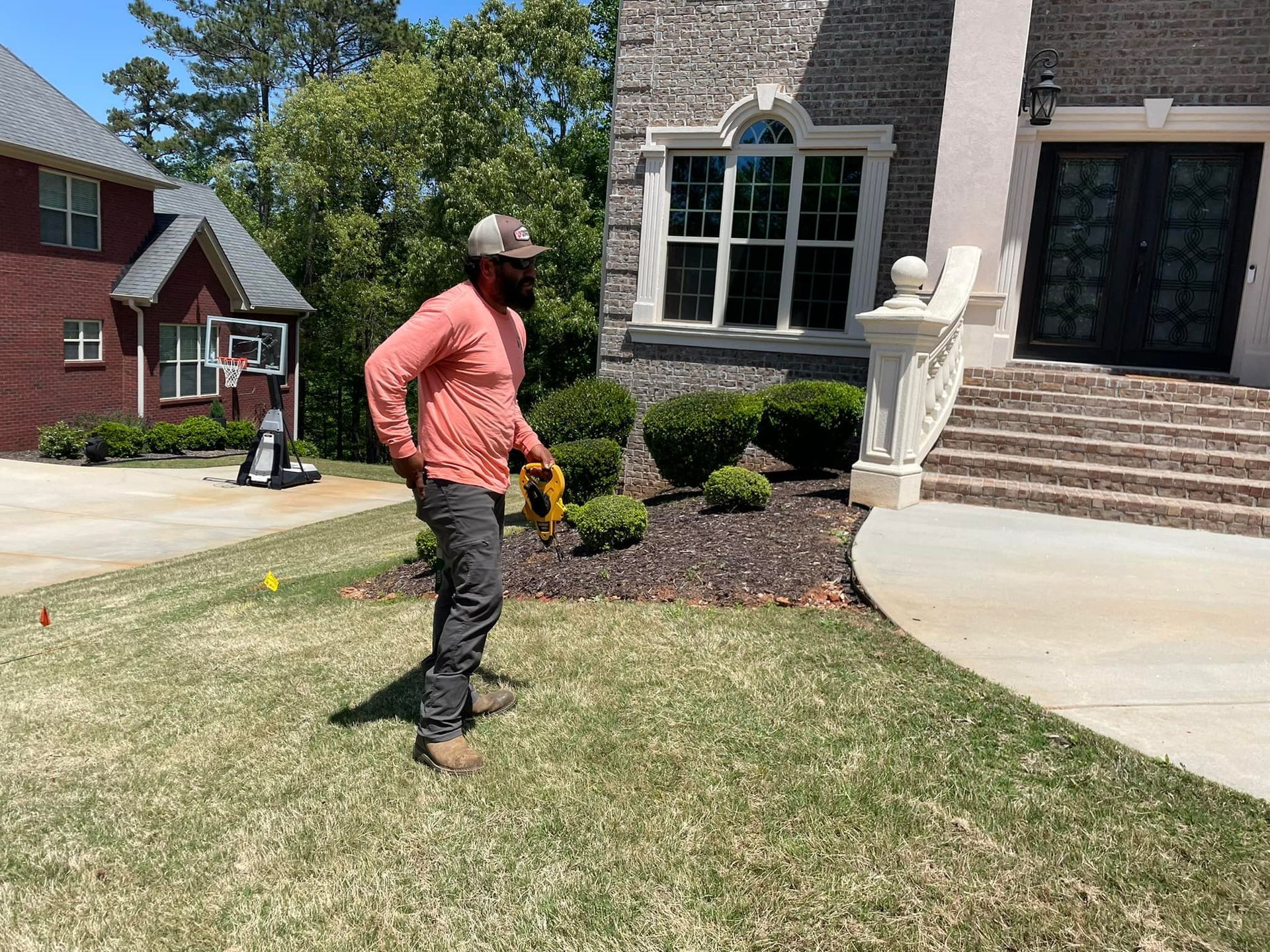 A person wearing a hat and long-sleeve shirt stands on grass near a house and garden bed.