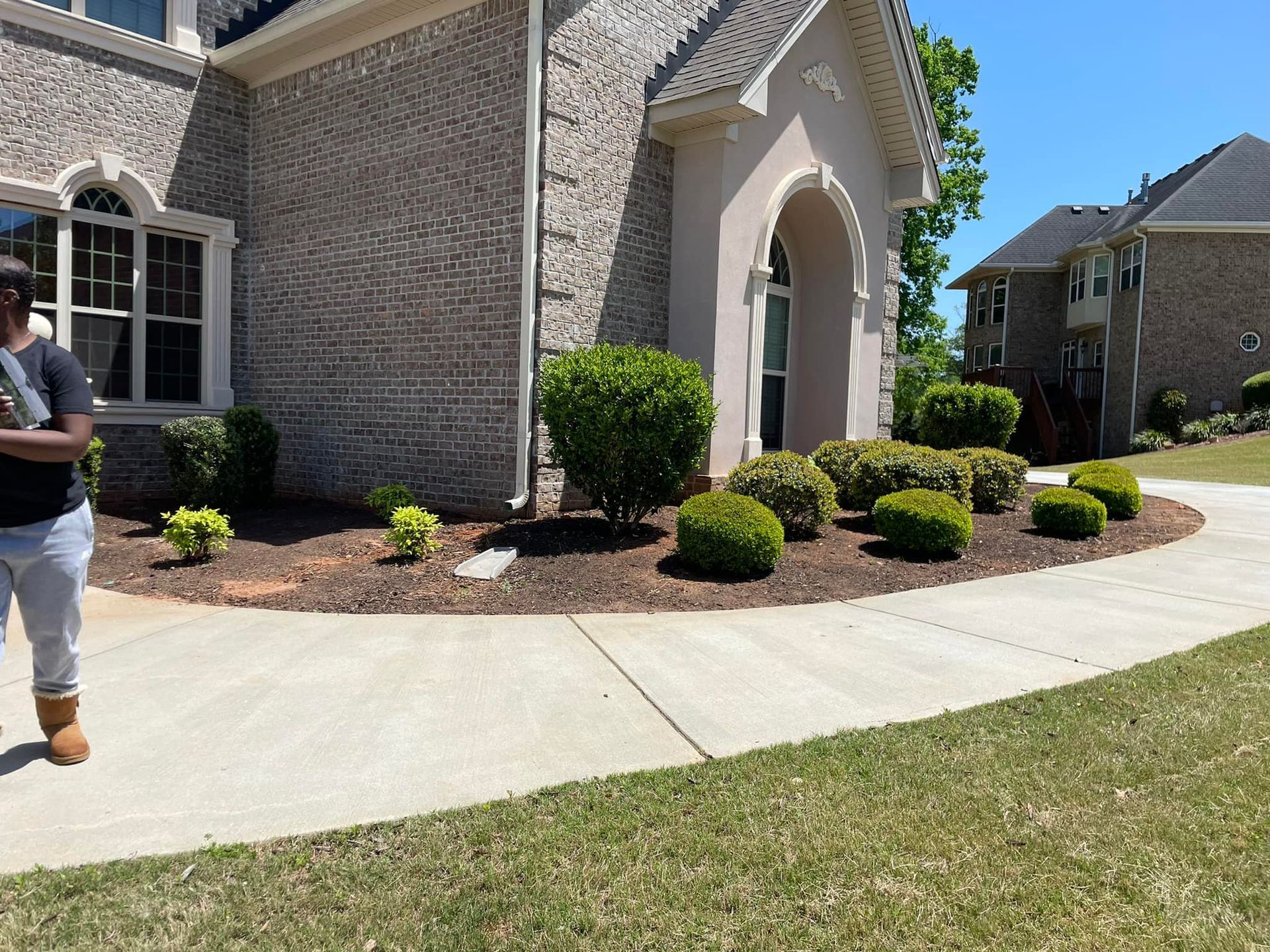 Person near a house with landscaping, a curved sidewalk, and neatly trimmed shrubs.