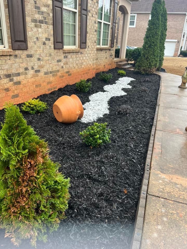 Front yard with black mulch, a rock river bed, and green bushes. An overturned terracotta pot rests near the plants.