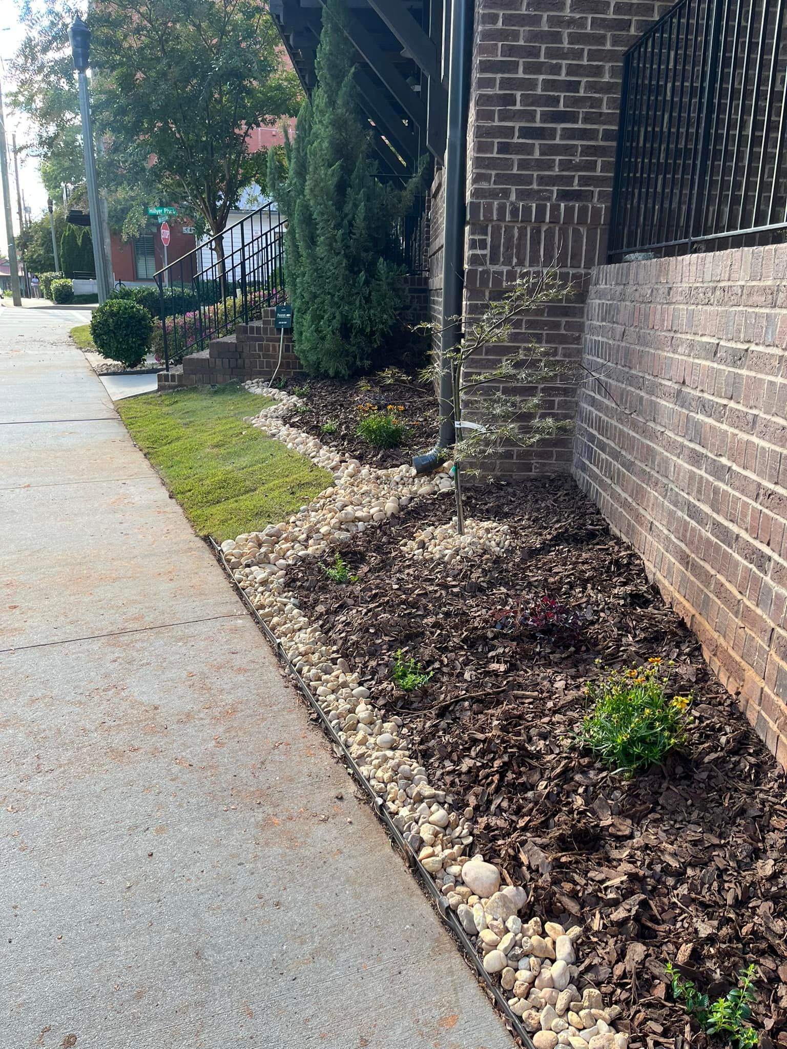 Sidewalk lined with landscaping: grass, mulch, and stone border next to a brick wall.