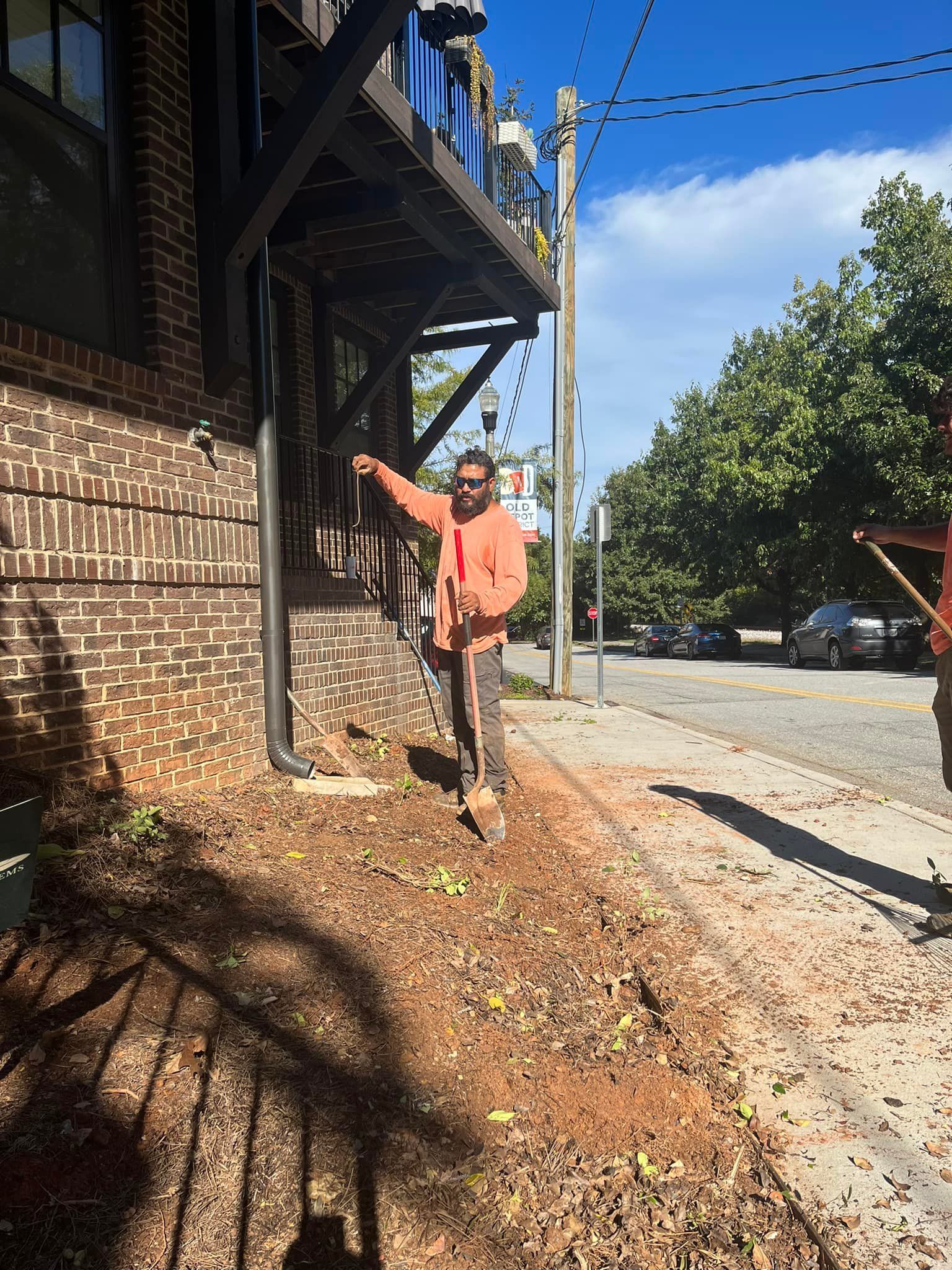 Man in orange shirt points near a building with bare earth and a street with trees.