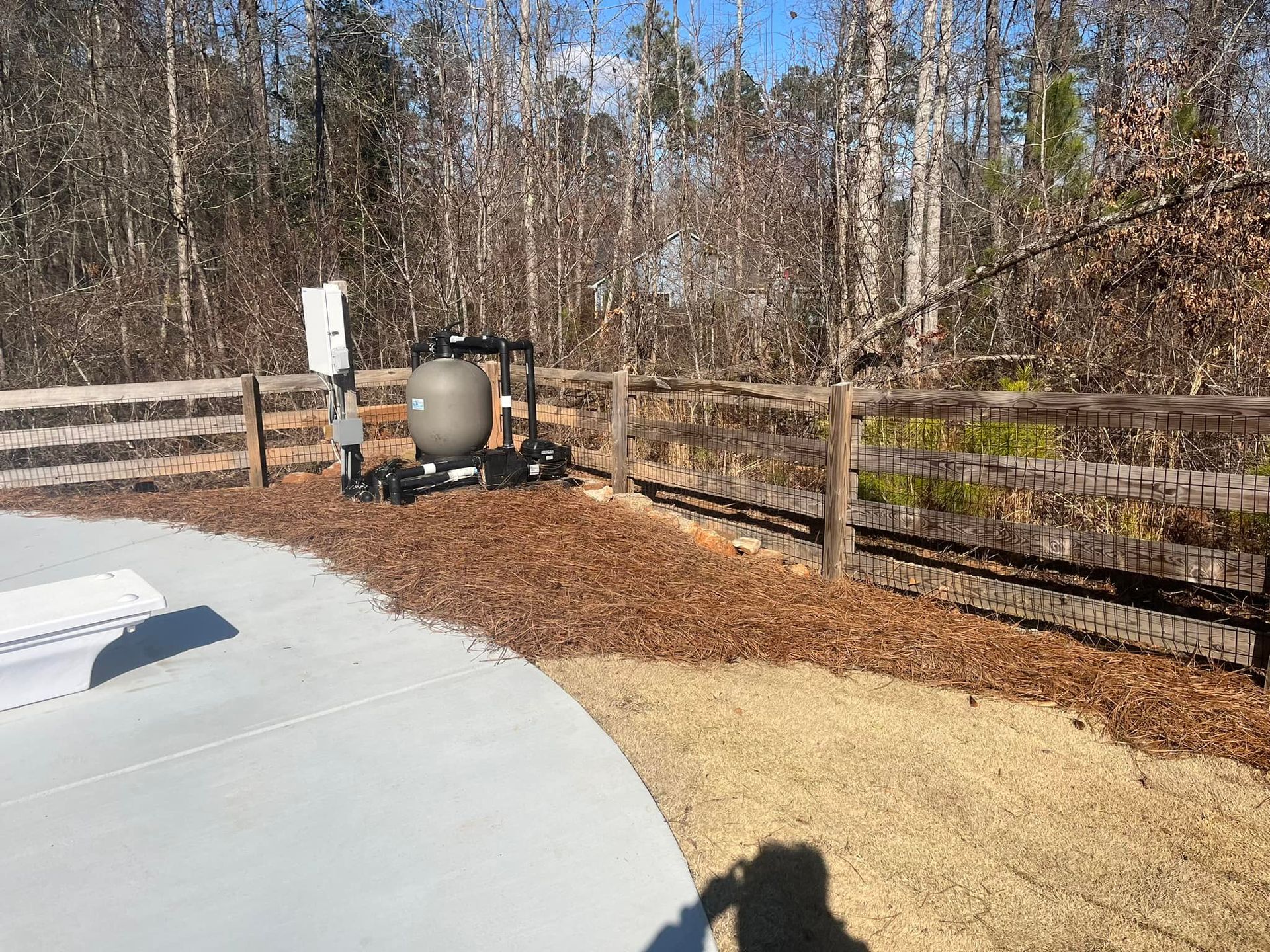Pool equipment next to a wooden fence and mulch. Bare trees in the background, concrete patio in the foreground.