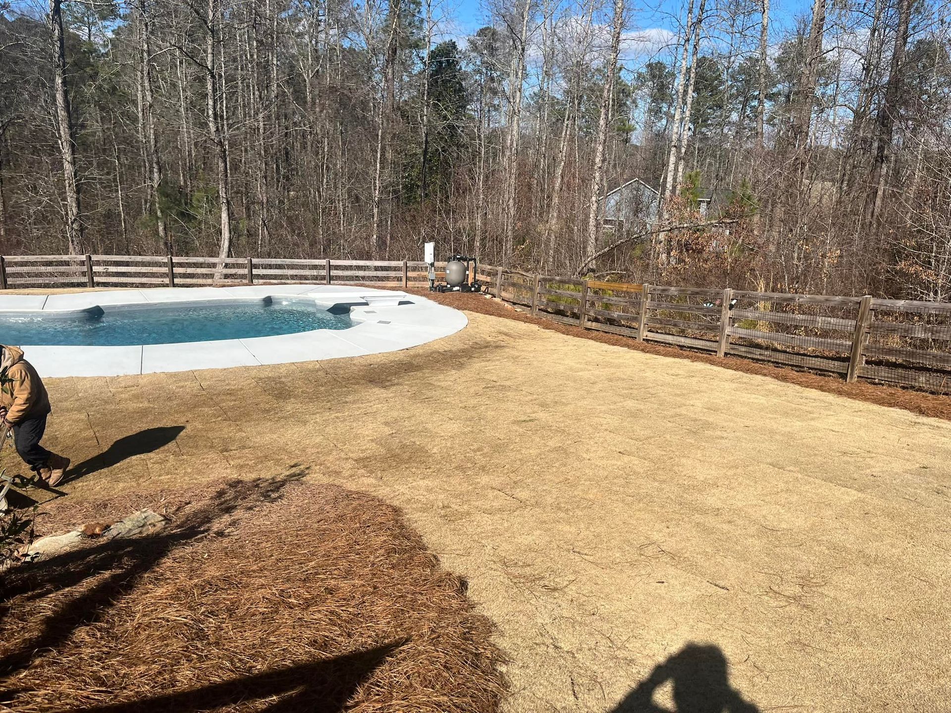 Backyard with pool, brown grass, wooden fence, person walking, and blue sky.