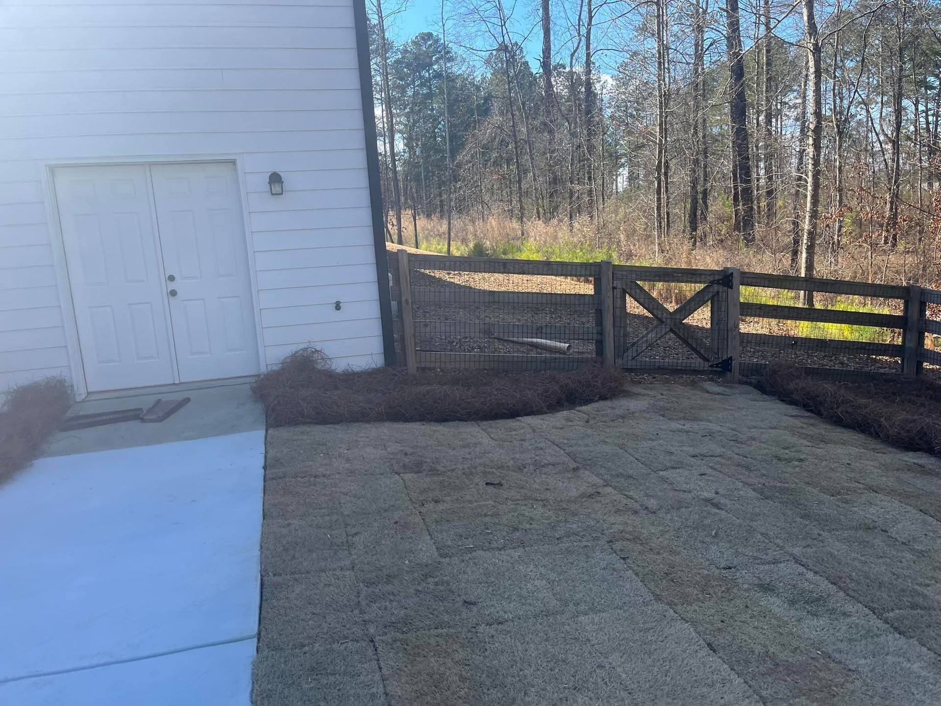 A white building with garage doors, concrete patio, and a wooden fence leading to a wooded area.
