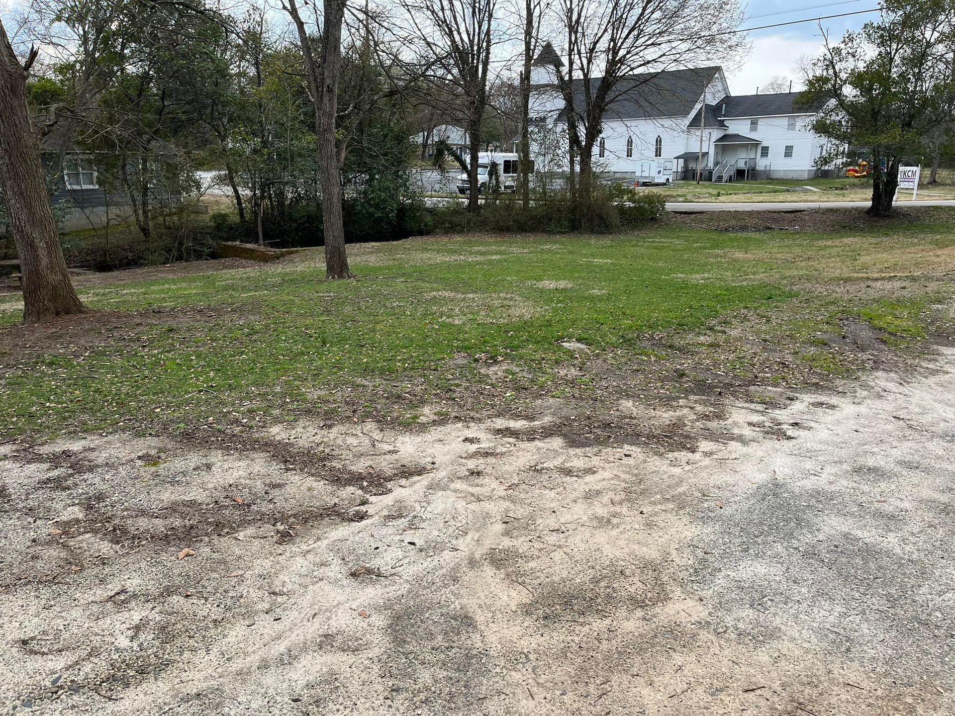 Grassy lot with trees in the foreground, white building in the background.