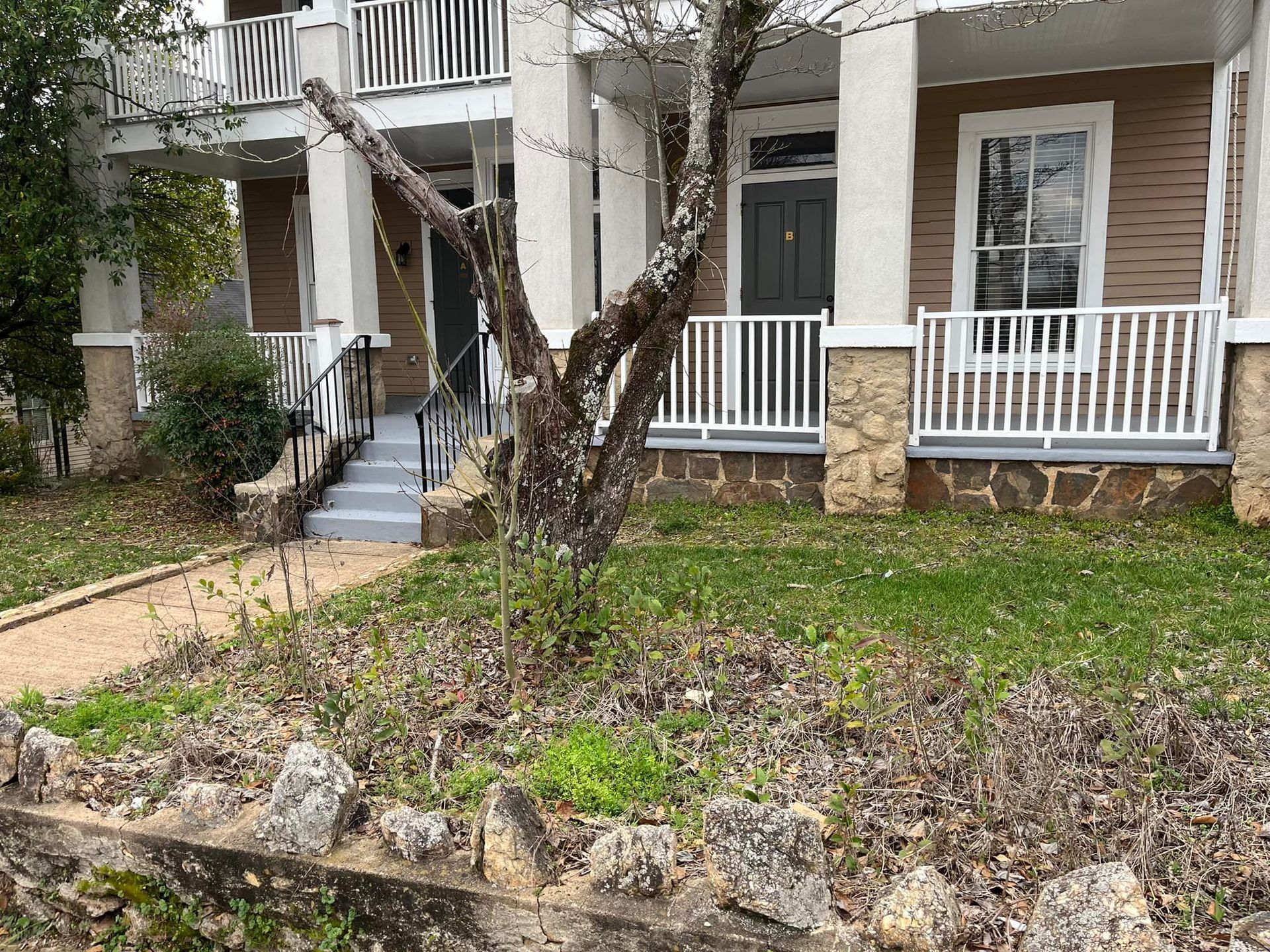 Two-story tan and white building with porch and balconies. A tree is in front. Stone fence and green grass.