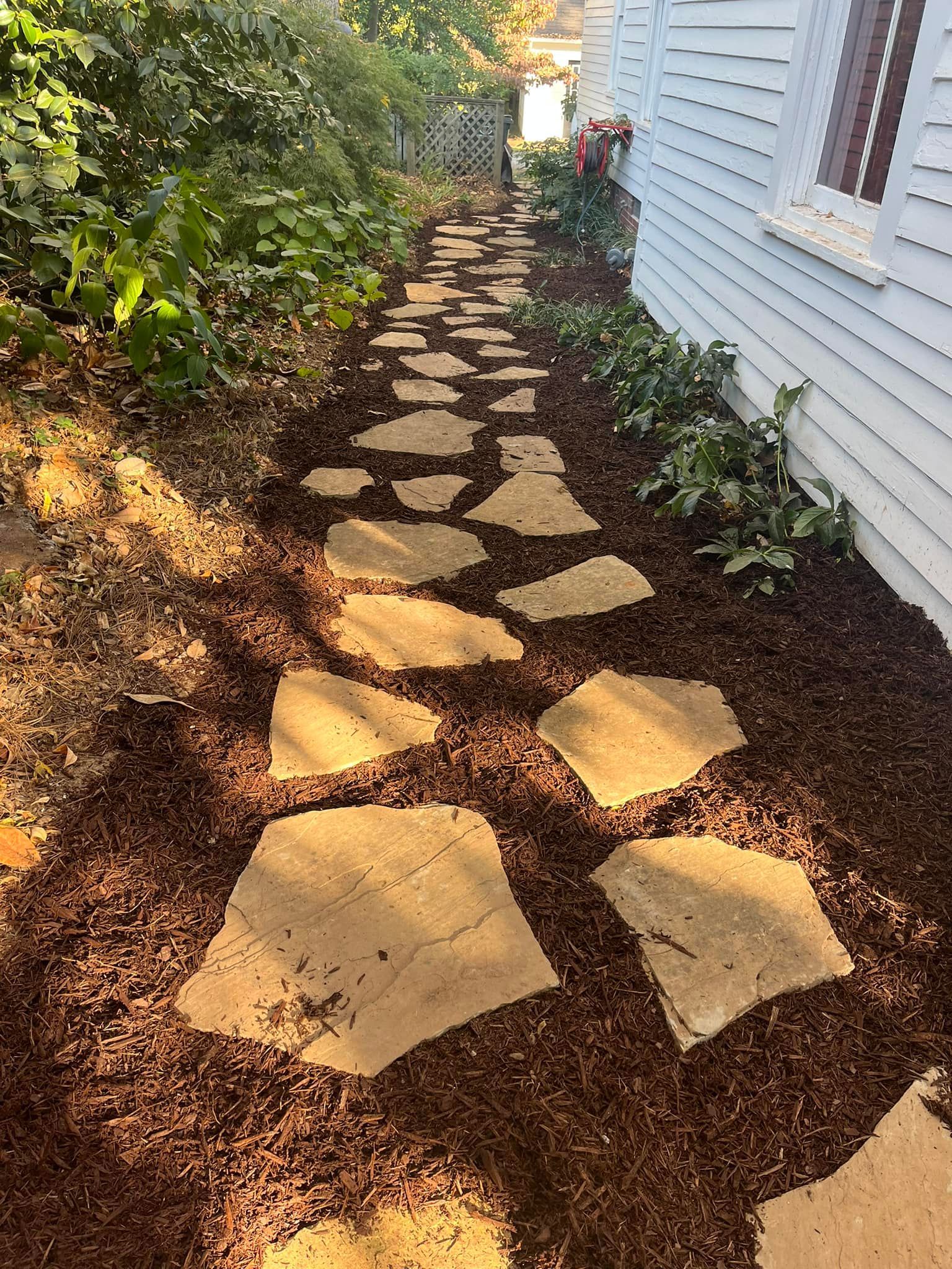 Stone pathway through a garden, lined with mulch and greenery, alongside a white house.