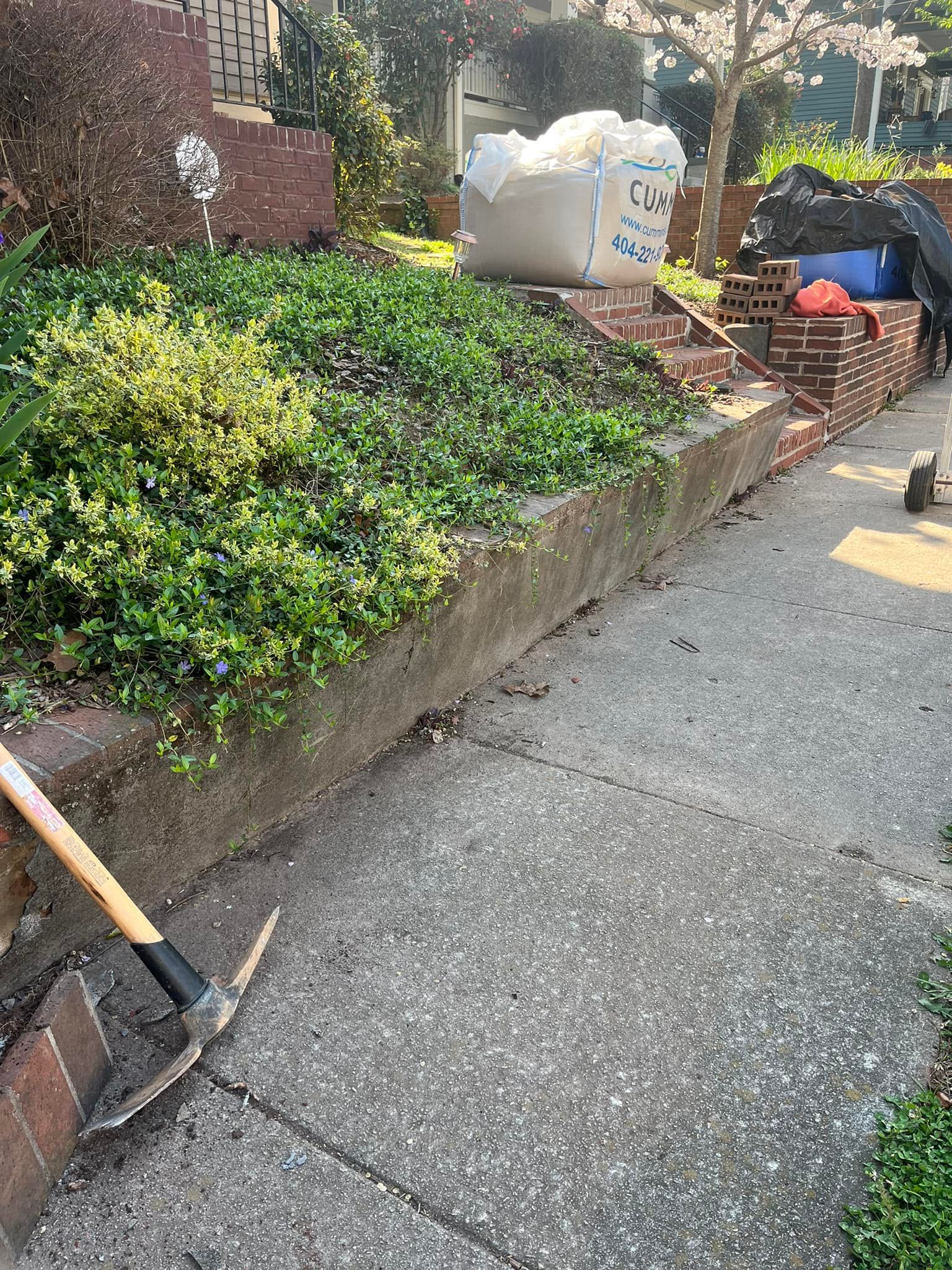 Concrete and brick retaining wall with plants, sidewalk, pickaxe, and a bag.