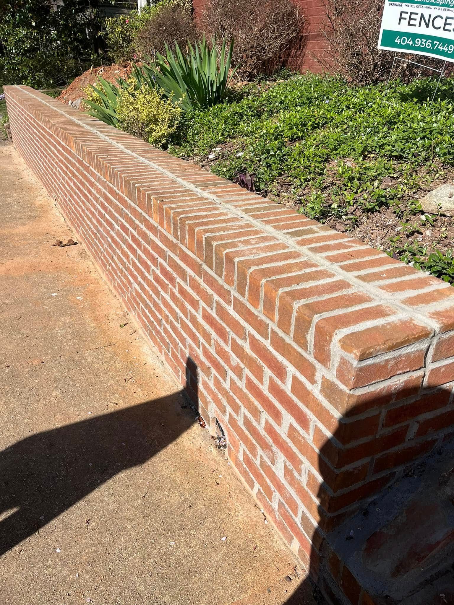 Brick retaining wall in front of greenery. Brown bricks with light mortar, next to a concrete walkway.
