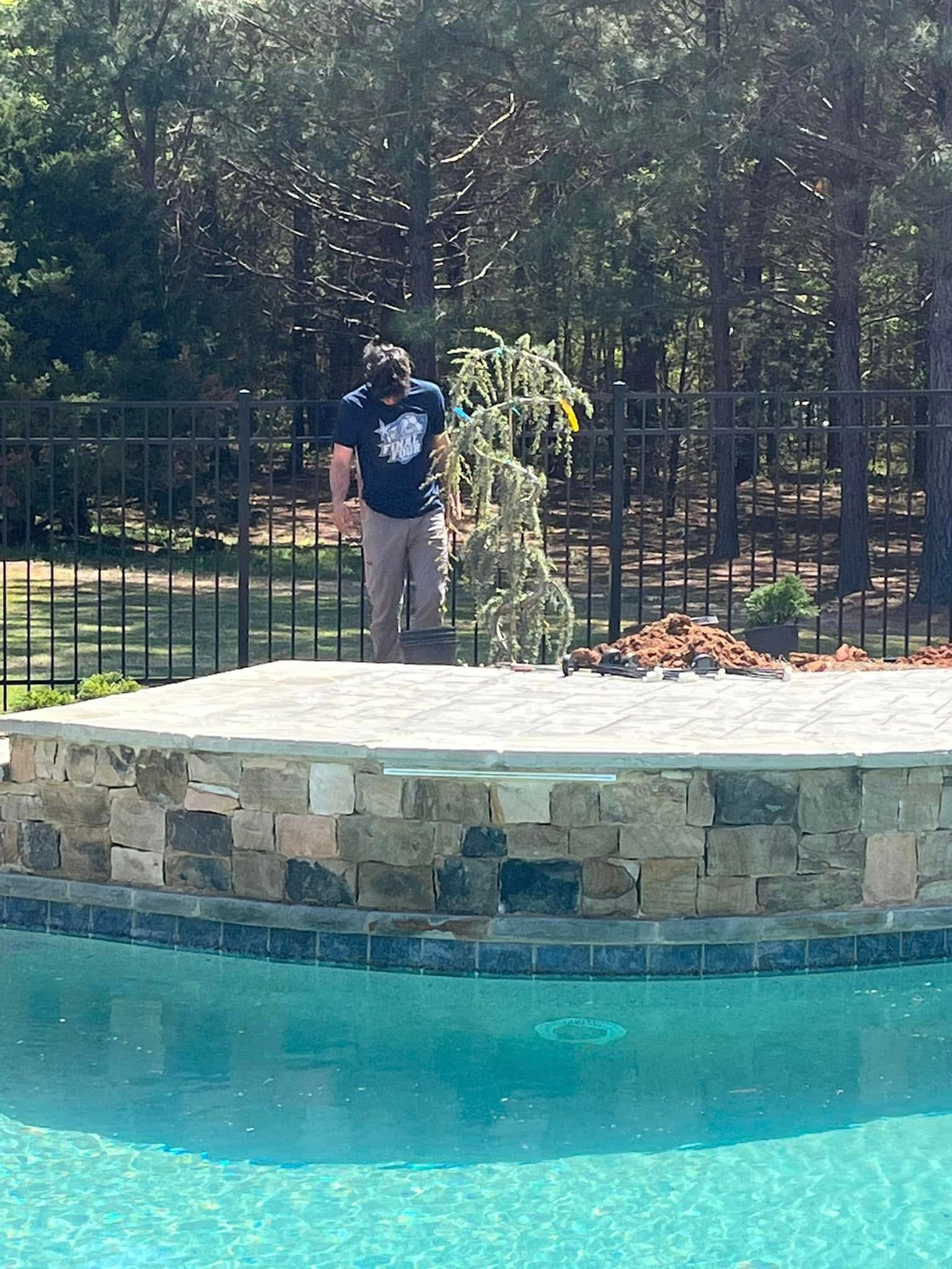 Person trimming a small tree on a stone patio by a pool, black fence in the background.