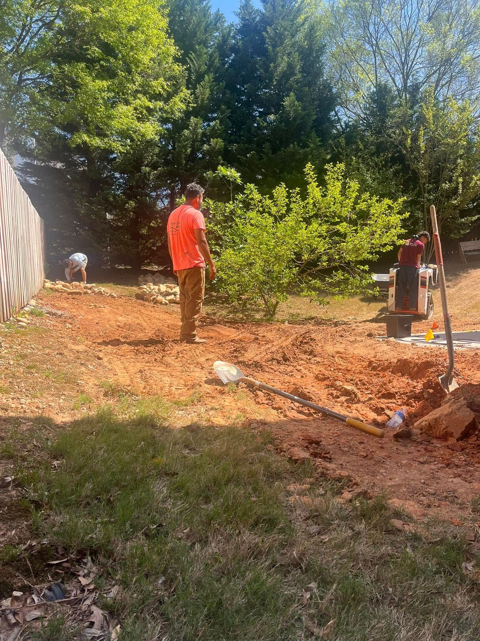 Workers in an orange shirt and tan pants preparing a yard for construction on a sunny day.